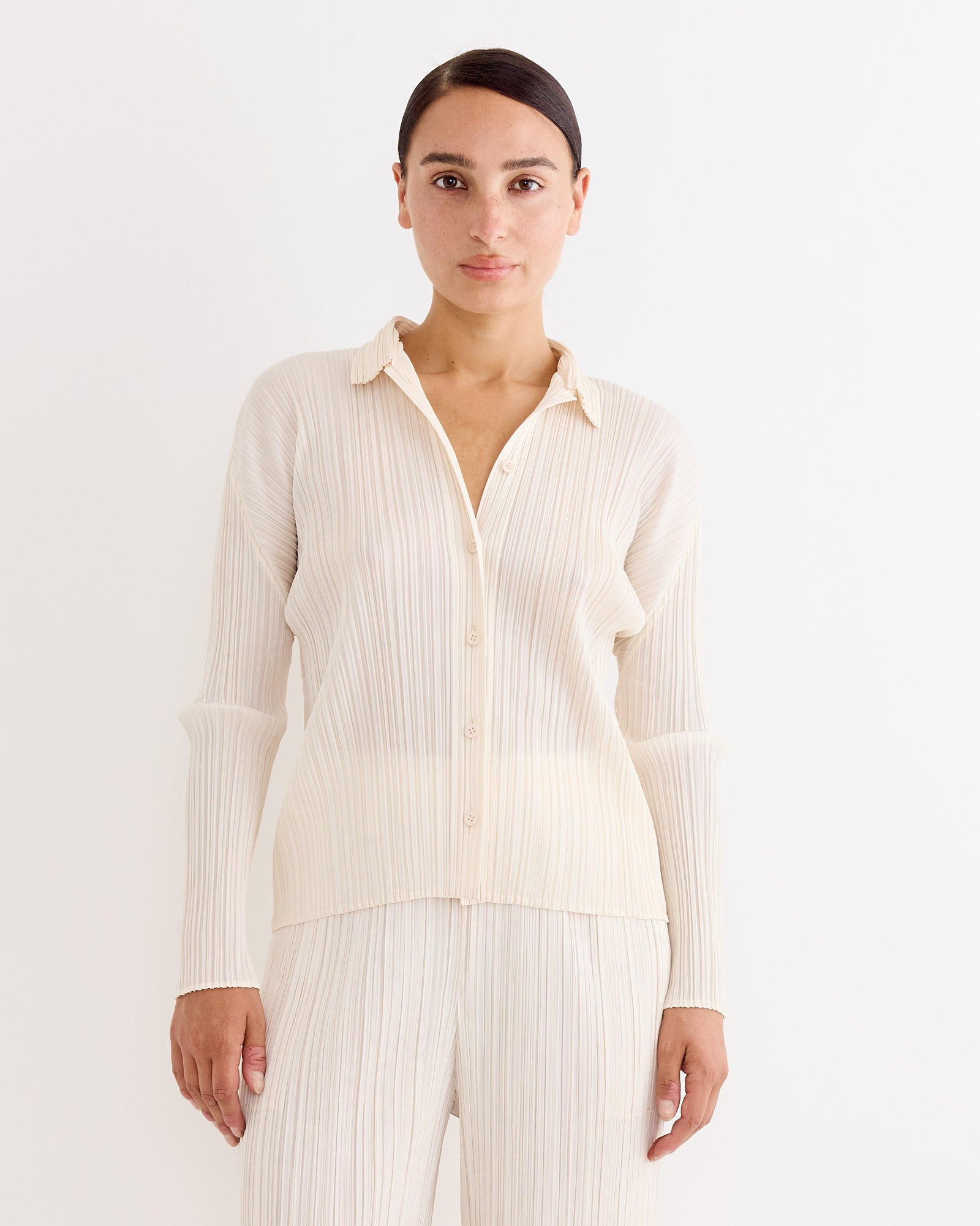 A person faces forward, wearing the Pleats Please Issey Miyake Shirt in Off White with subtle vertical stripes and matching pants, set against a plain white background.