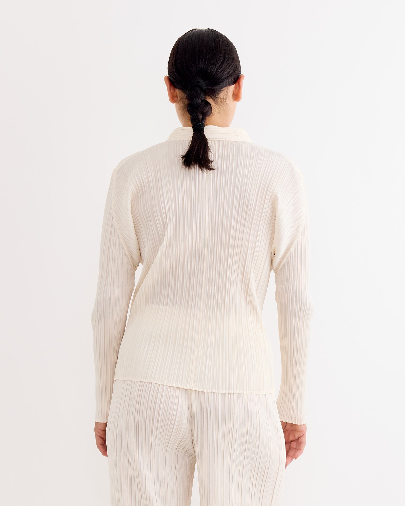 A person with dark hair in a low braid, wearing the Pleats Please Issey Miyake Shirt in Off White, stands facing away against a plain white background.