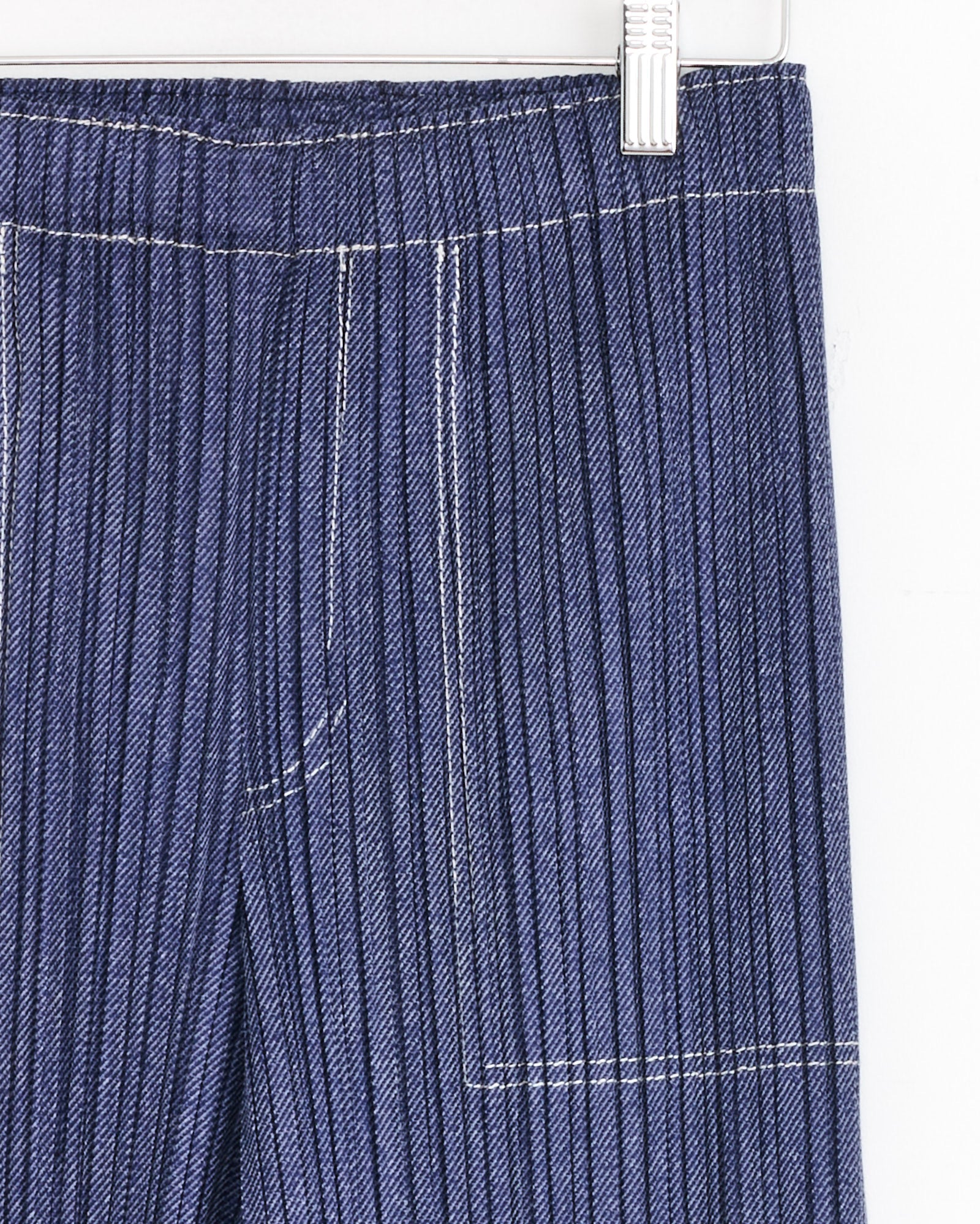 Close-up of PLEATS PLEASE ISSEY MIYAKE's Cotton Denim in Blue pants with vertical white pinstripes, a large front patch pocket, white stitching, and a metal hanger clip at the waistband.