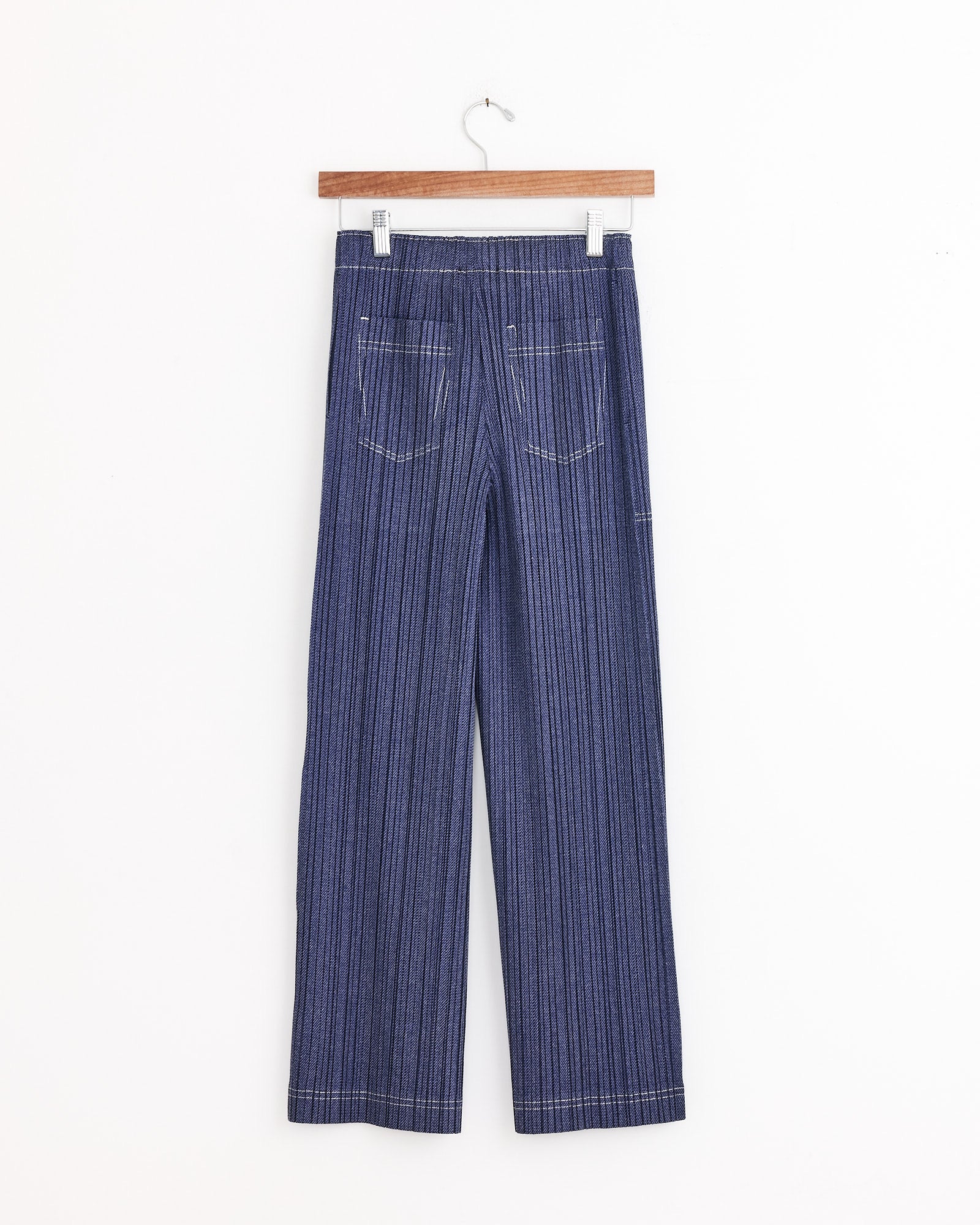 The Cotton Denim in Blue by PLEATS PLEASE ISSEY MIYAKE are blue, pinstriped wide-leg pants with an elastic waistband and two back pockets, displayed on a wooden hanger against a white background.