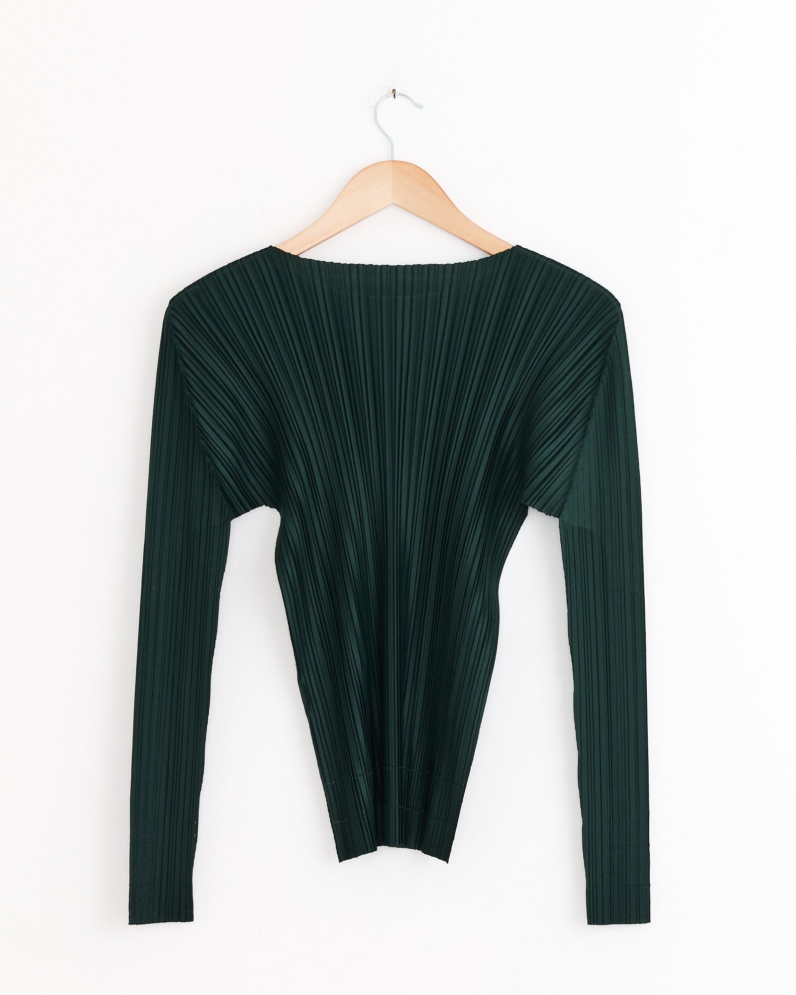 The Soft Top in Dark Green by PLEATS PLEASE ISSEY MIYAKE, featuring long sleeves and a boat neckline, hangs on a wooden hanger against a plain white wall.