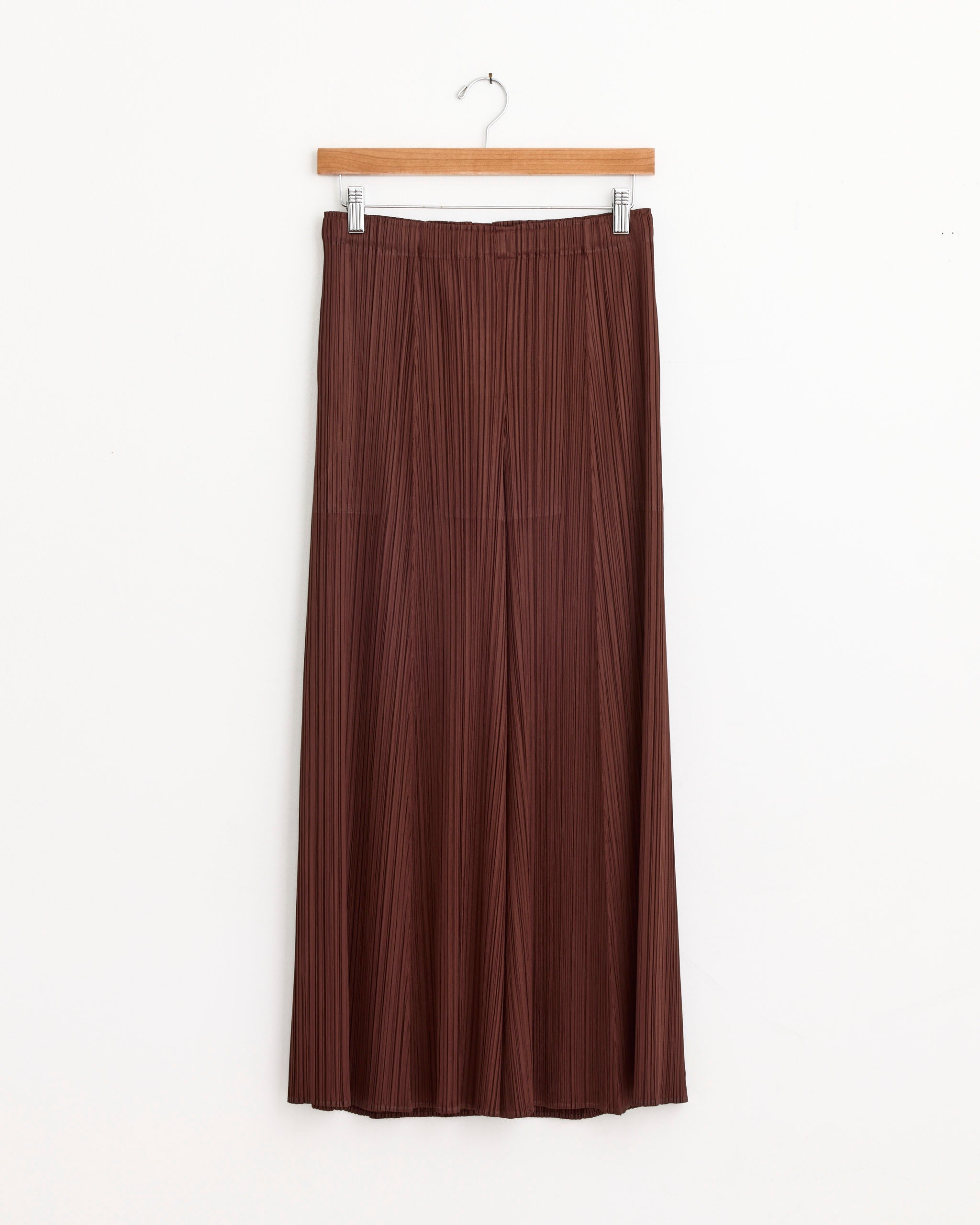The Nov Pant in Brown from Pleats Please Issey Miyake hangs on a wooden hanger against a plain white background. For fit or sizing questions, contact our online chat for assistance.