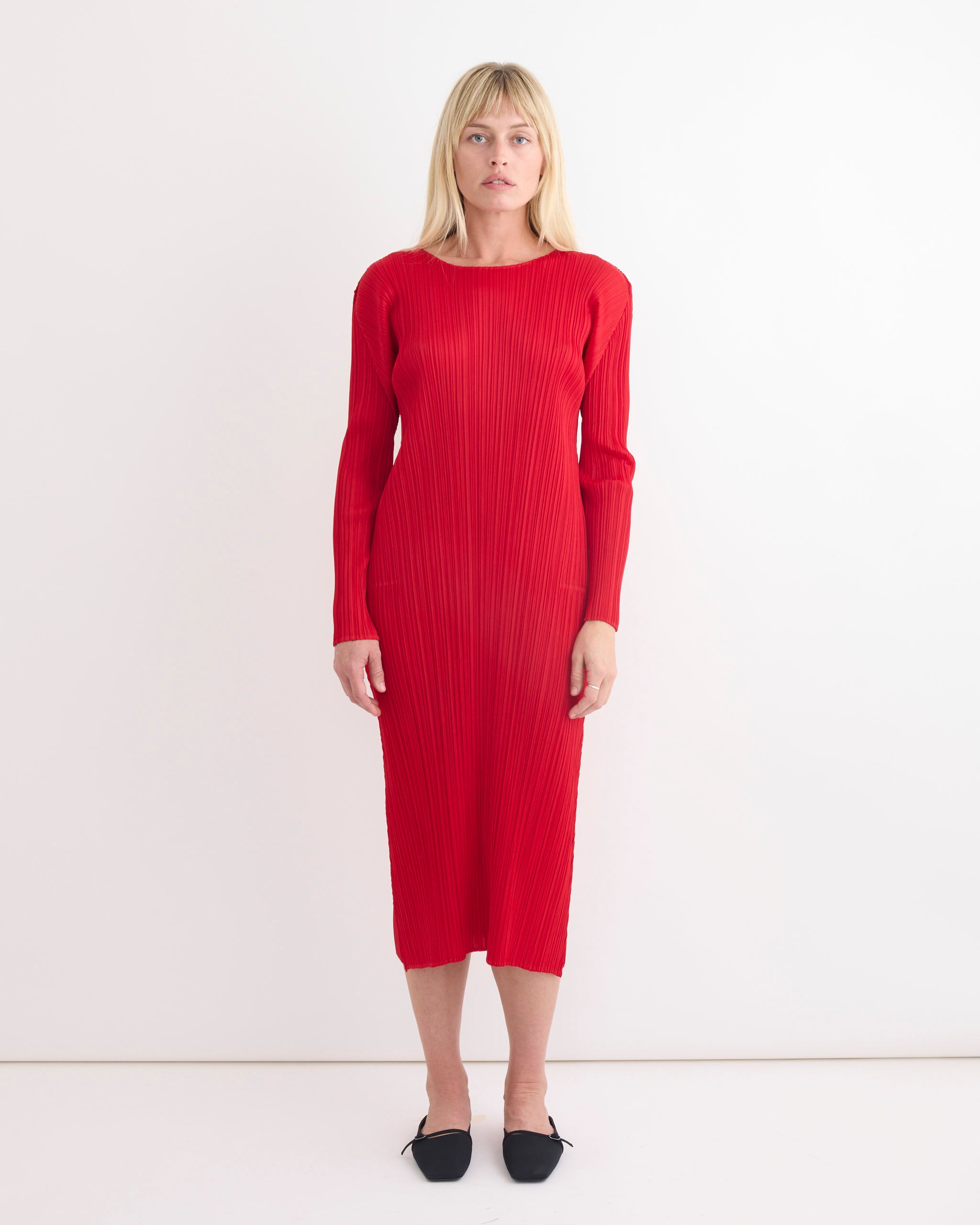 A woman with straight blonde hair models the Pleats Please Issey Miyake MC Nov Dress in Red—a long-sleeved, ribbed midi dress—paired with black pointed-toe flats, standing against a plain white background to highlight fit and sizing.