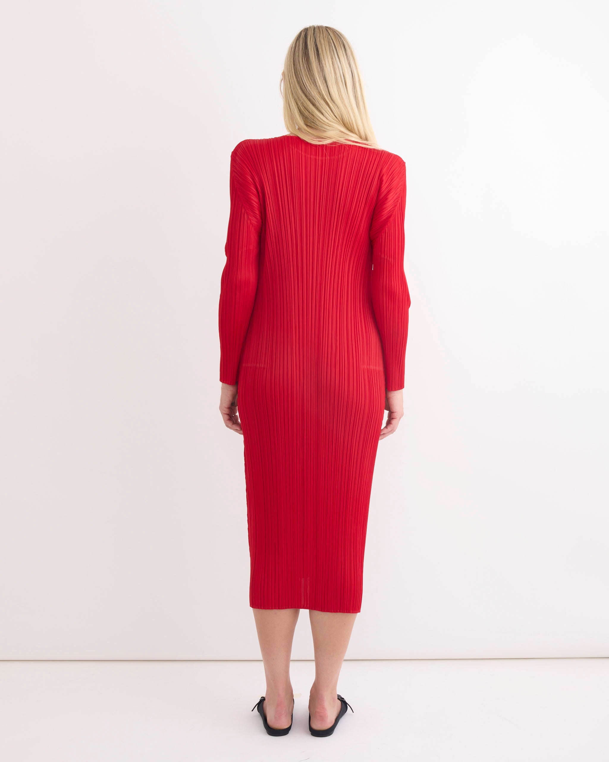A woman with long blonde hair faces away, modeling the Pleats Please Issey Miyake MC Nov Dress in Red—fitted, knee-length, long-sleeved with vertical pleats—paired with black heels against a white backdrop for online sizing checks.