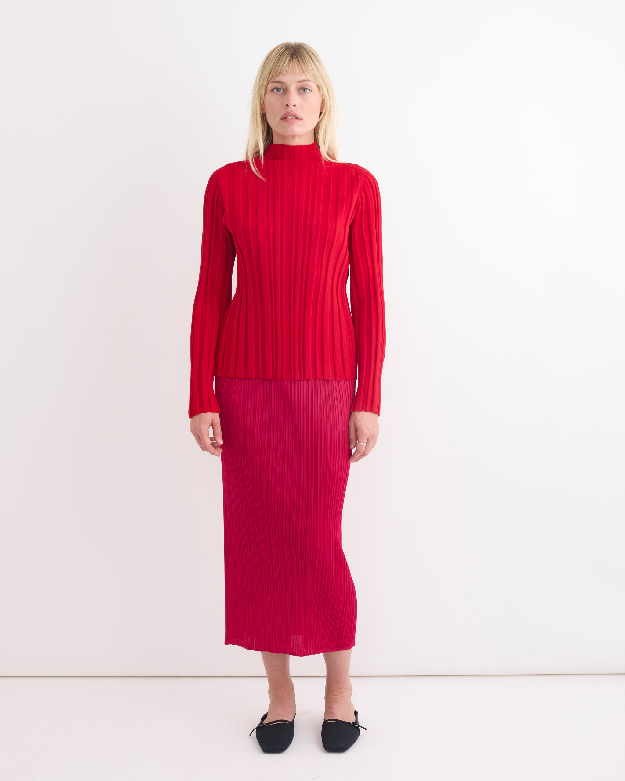 A woman in the Rib Knit in Red by Pleats Please Issey Miyake stands before a plain white wall, wearing the long-sleeved top and matching midi skirt, with straight blonde hair and black pointed-toe flats.