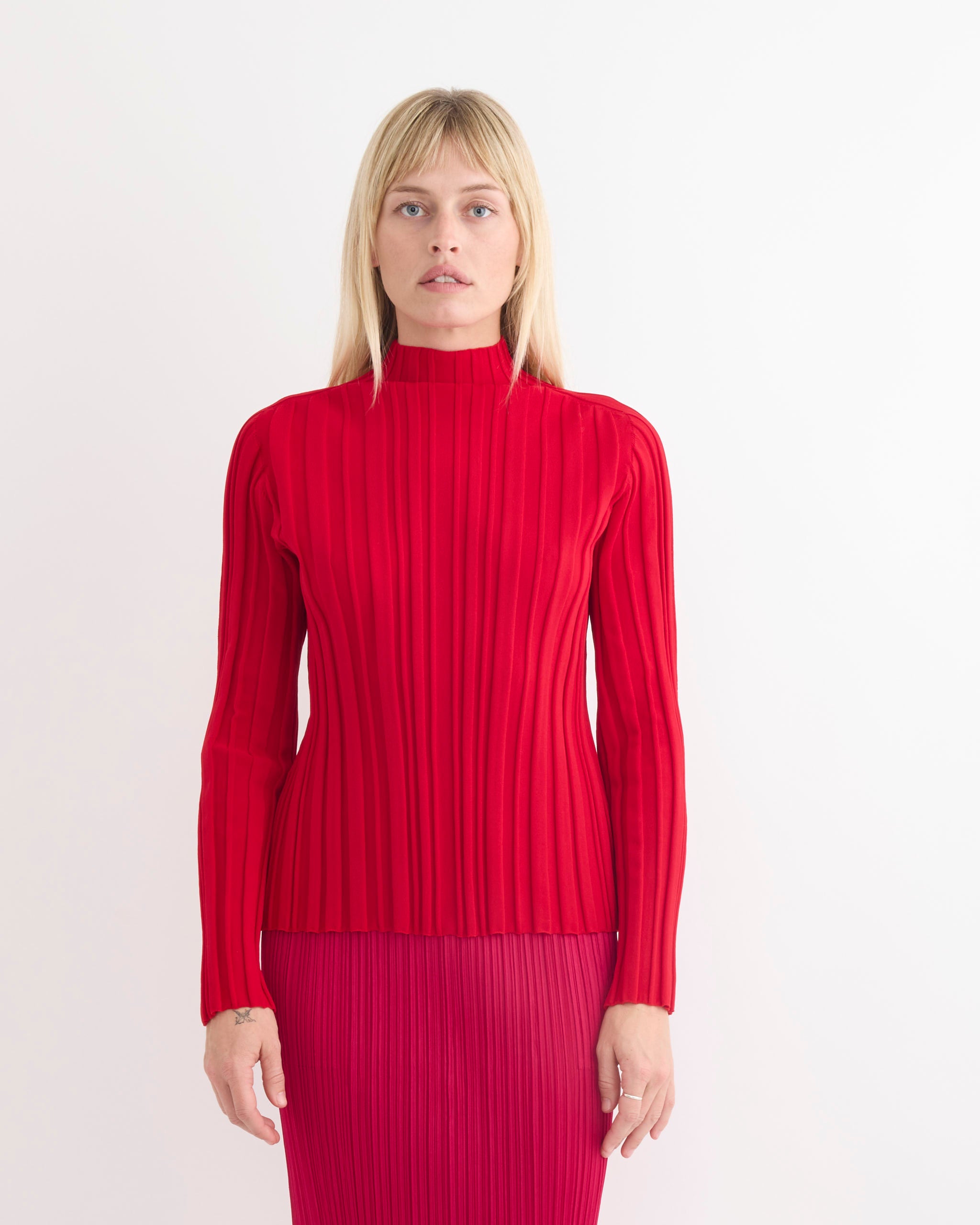 A woman with straight blonde hair wears the Pleats Please Issey Miyake Rib Knit in Red, featuring long sleeves and a high neck, paired with a pleated red skirt, standing against a plain white background.