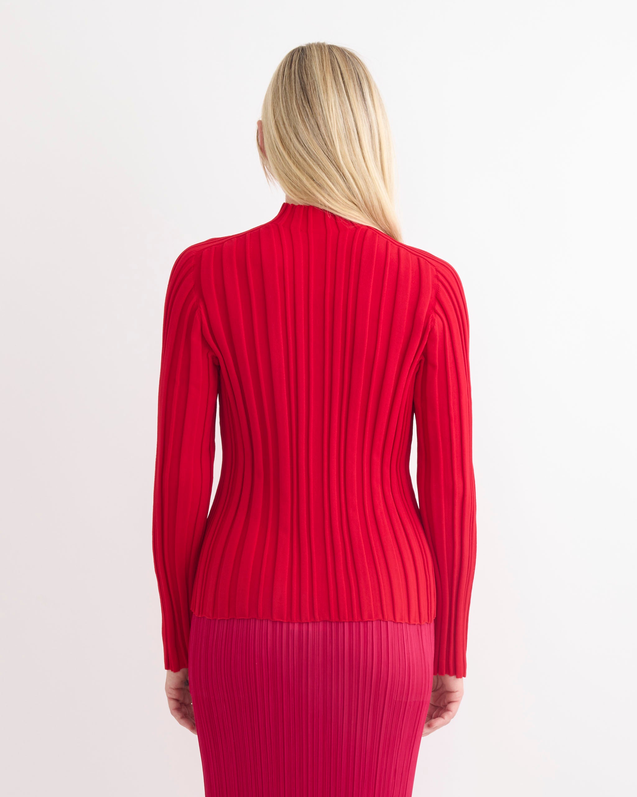 A person with long blonde hair, seen from behind, wears the Rib Knit in Red set by Pleats Please Issey Miyake—a fitted, long-sleeved rib knit top and matching skirt—against a plain white background.