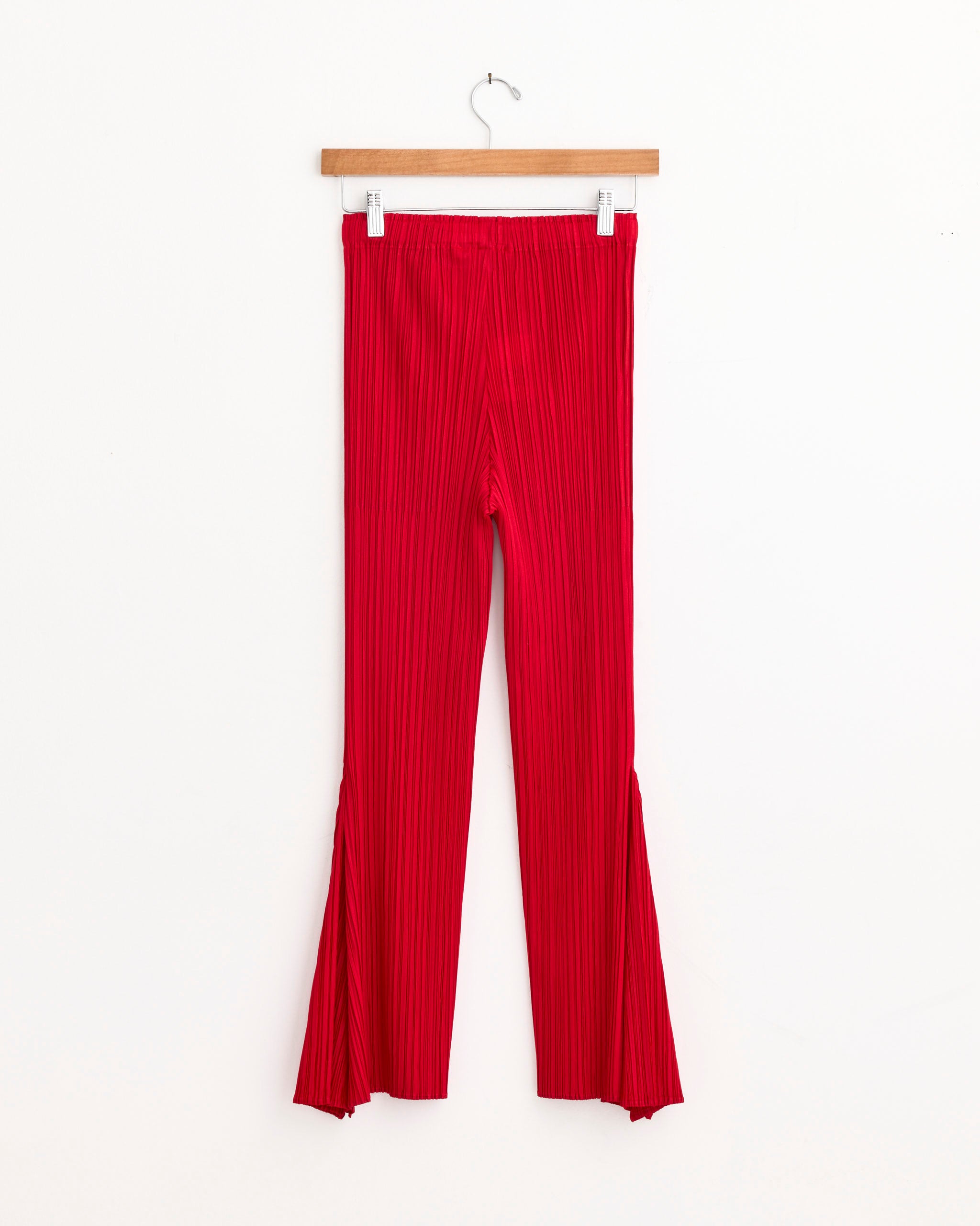 The Pleats Please Issey Miyake Ole! Pant in Red, featuring bright pleats and flared slit hems, hangs on a wooden hanger against a white background. For fit or sizing questions, connect with our online chat team.