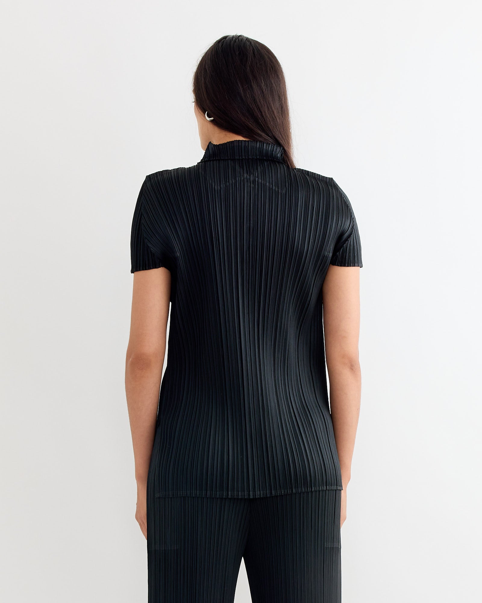 A person with long dark hair, facing away from the camera against a plain light background, wears the Pleats Please Issey Miyake MC August Top in Black with matching pants.
