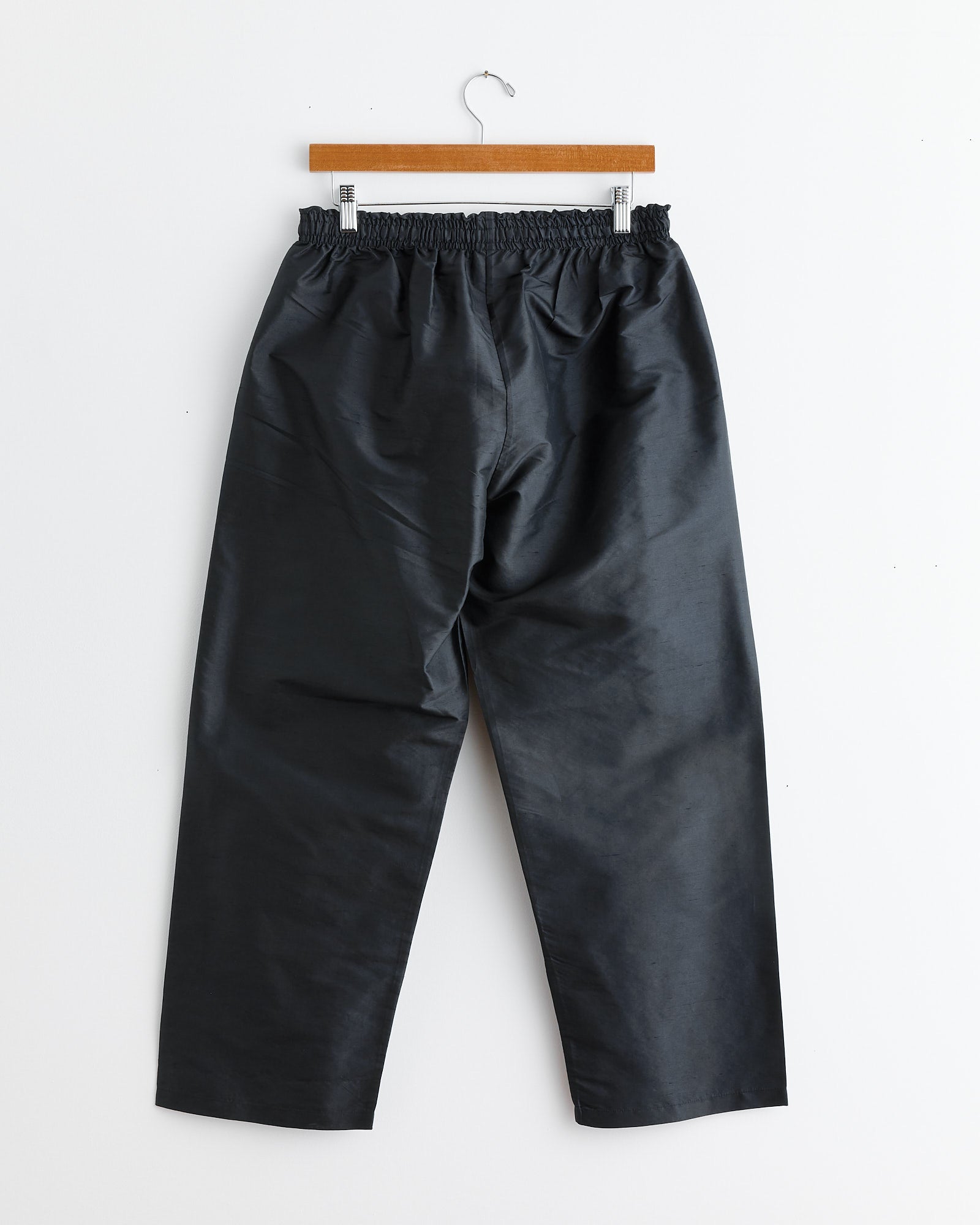 The Lexy Pant in Black by Chelsea Mak, crafted from 100% Silk Taffeta with an elastic waist, hangs on a wooden hanger against a plain white wall.