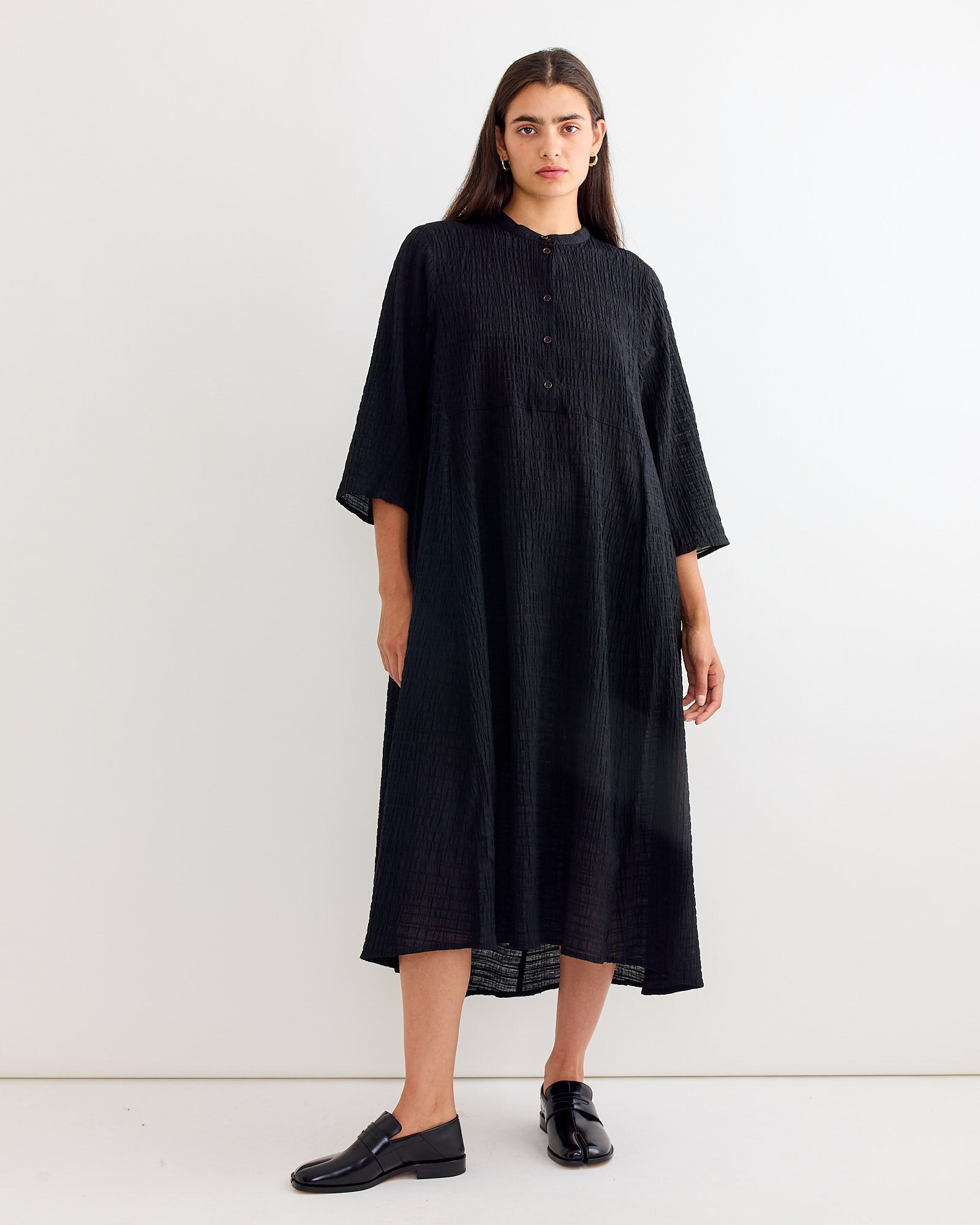 A woman with long dark hair stands against a white wall, wearing Henrik Vibskov’s Low Pulse Dress in Black—a loose, textured dress with an asymmetrical hemline, elbow-length sleeves, and top buttons—paired with black loafers.