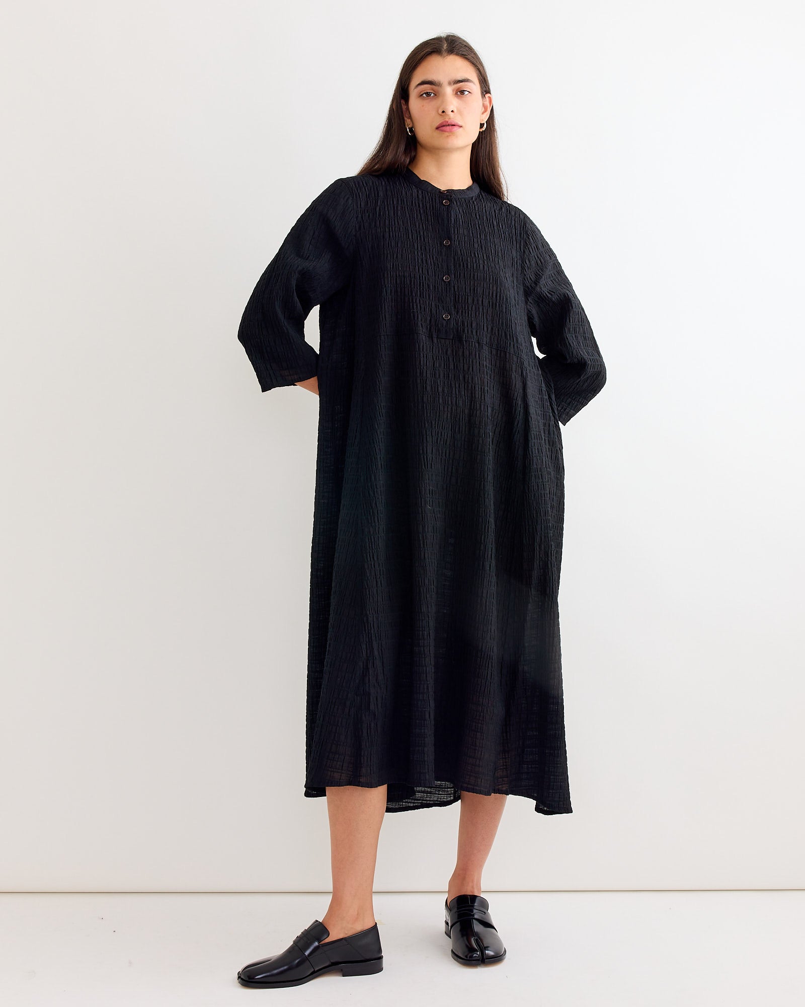 A person with long dark hair and a neutral expression stands against a white wall, wearing Henrik Vibskov's Low Pulse Dress in Black—featuring textured fabric, an asymmetrical hemline, long sleeves, front buttons—and black loafers.