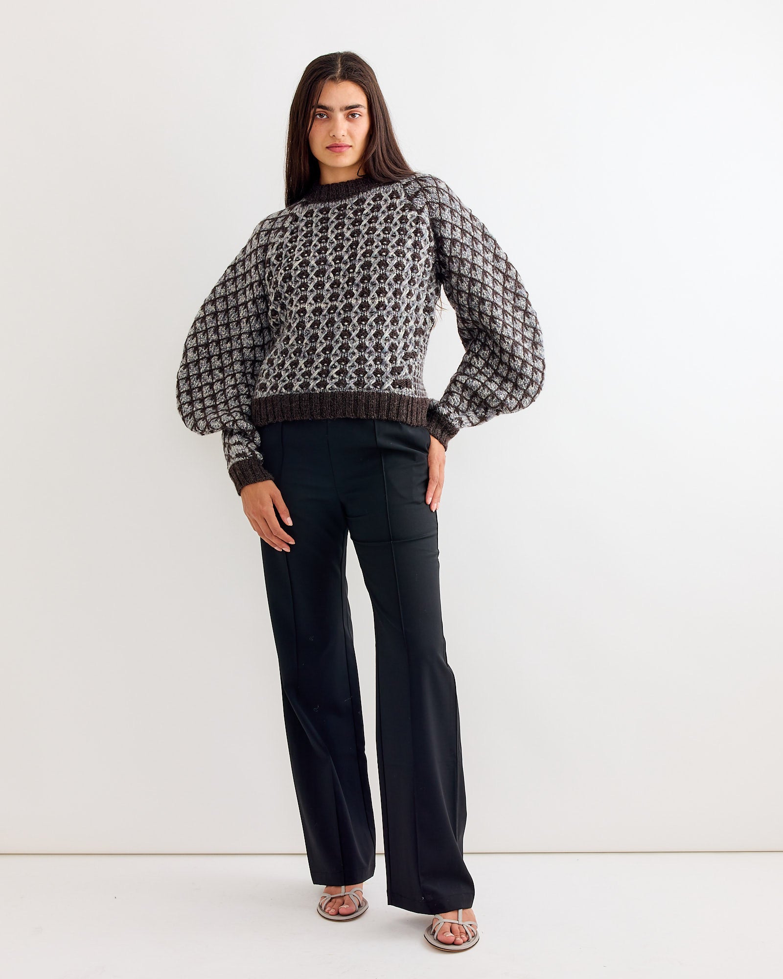 A woman with long dark hair wears the Henrik Vibskov Kimchi Sweater in Brown Party, paired with black pants and silver sandals, standing confidently against a plain white background.