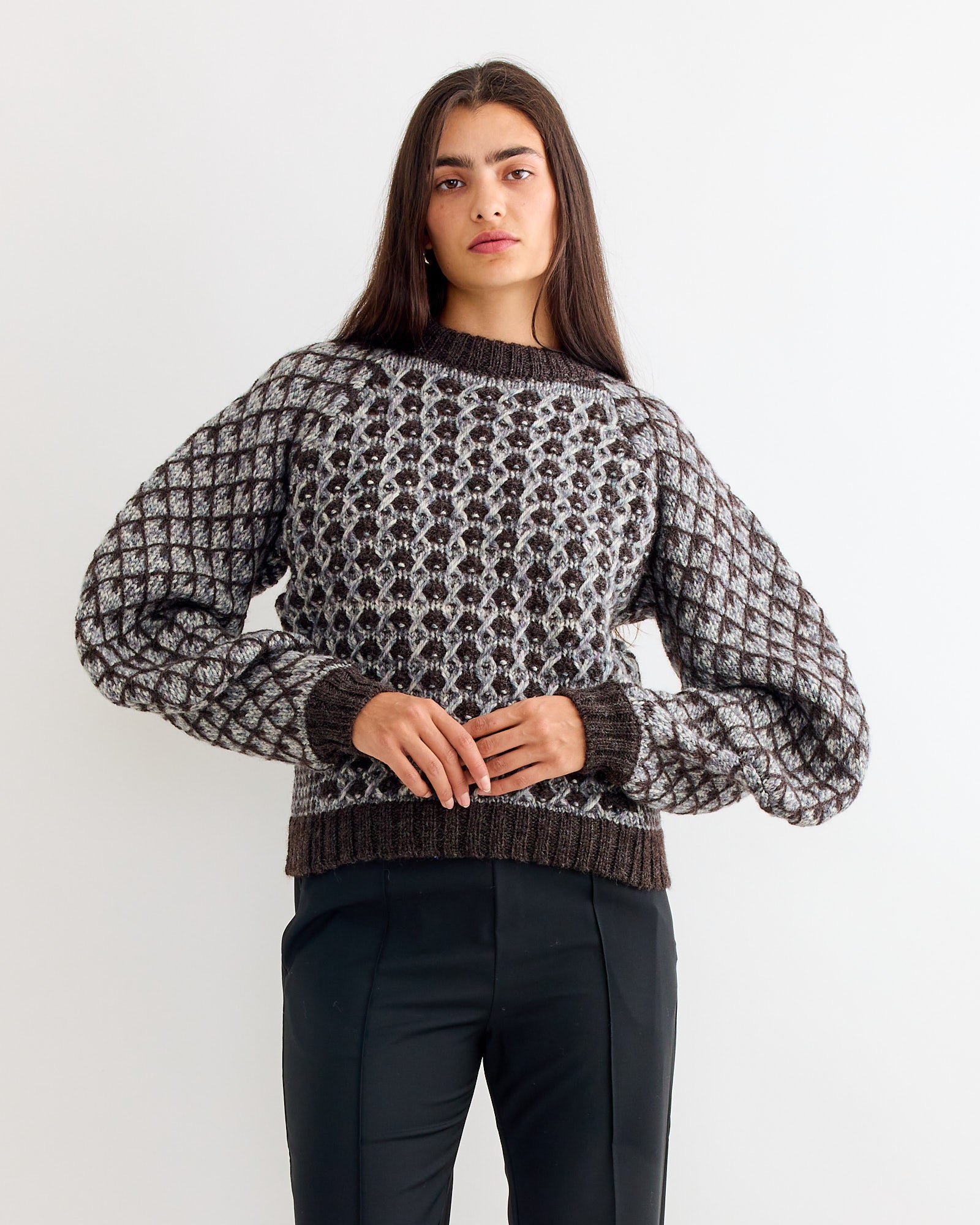 A woman with long dark hair wears the Henrik Vibskov Kimchi Sweater in Brown Party with black pants, standing against a plain white background.