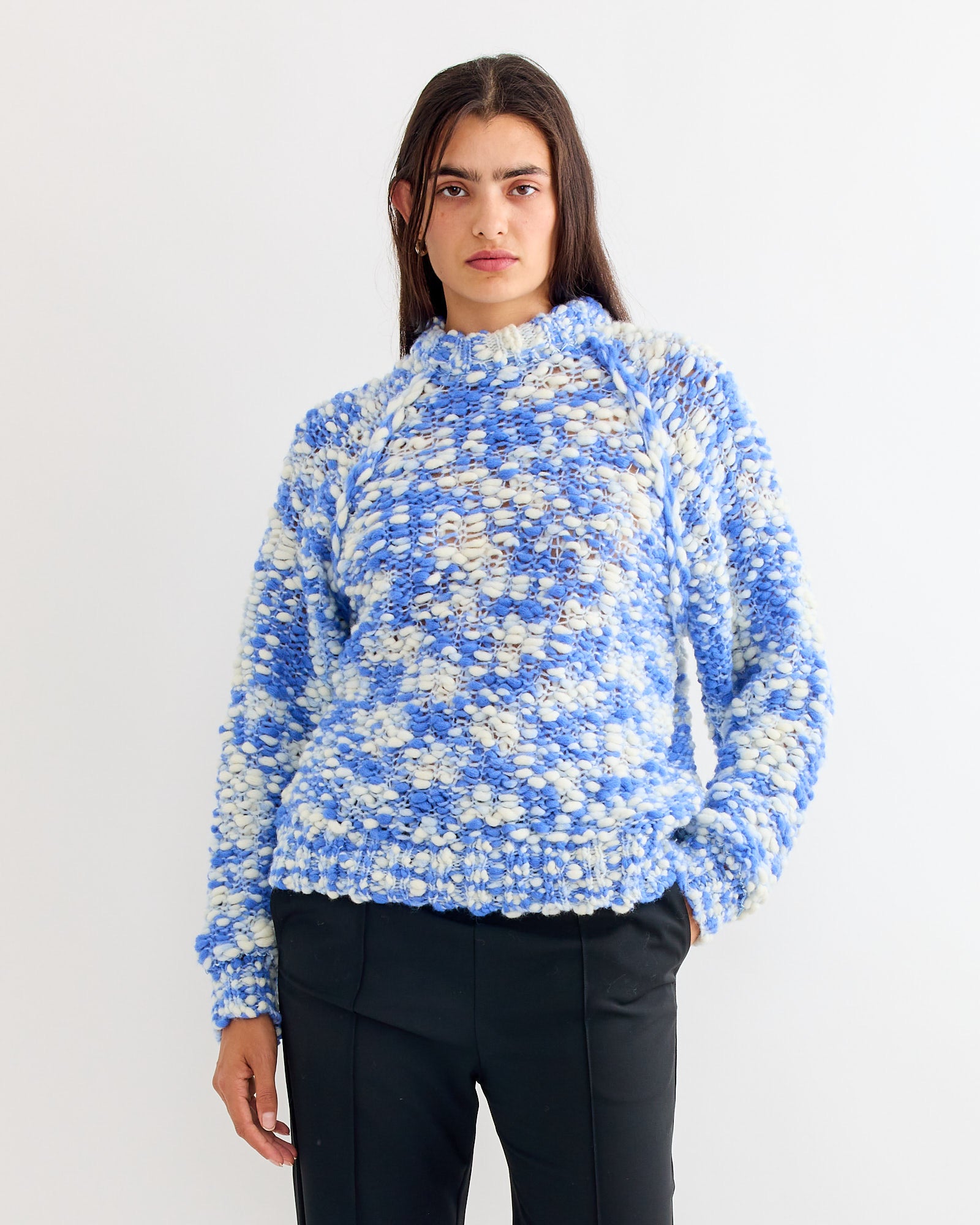 A person with long dark hair wears the Henrik Vibskov Bongo Sweater in Blue, styled with black pants, standing in front of a plain white background.
