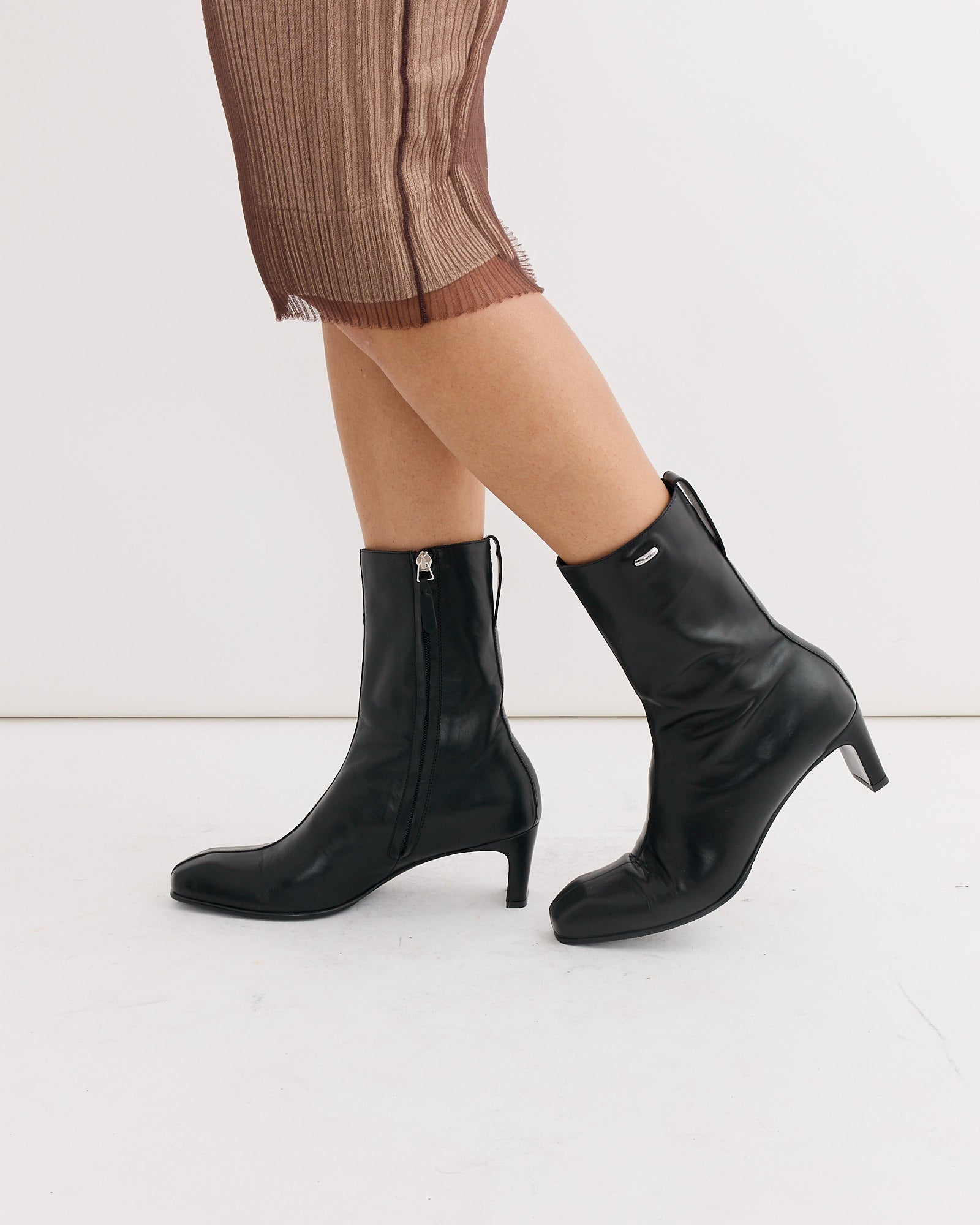 A person wearing a pleated brown skirt and black leather heeled boots, such as the Karasu Heel Boot in Black by Our Legacy with side zippers, stands against a plain white background.