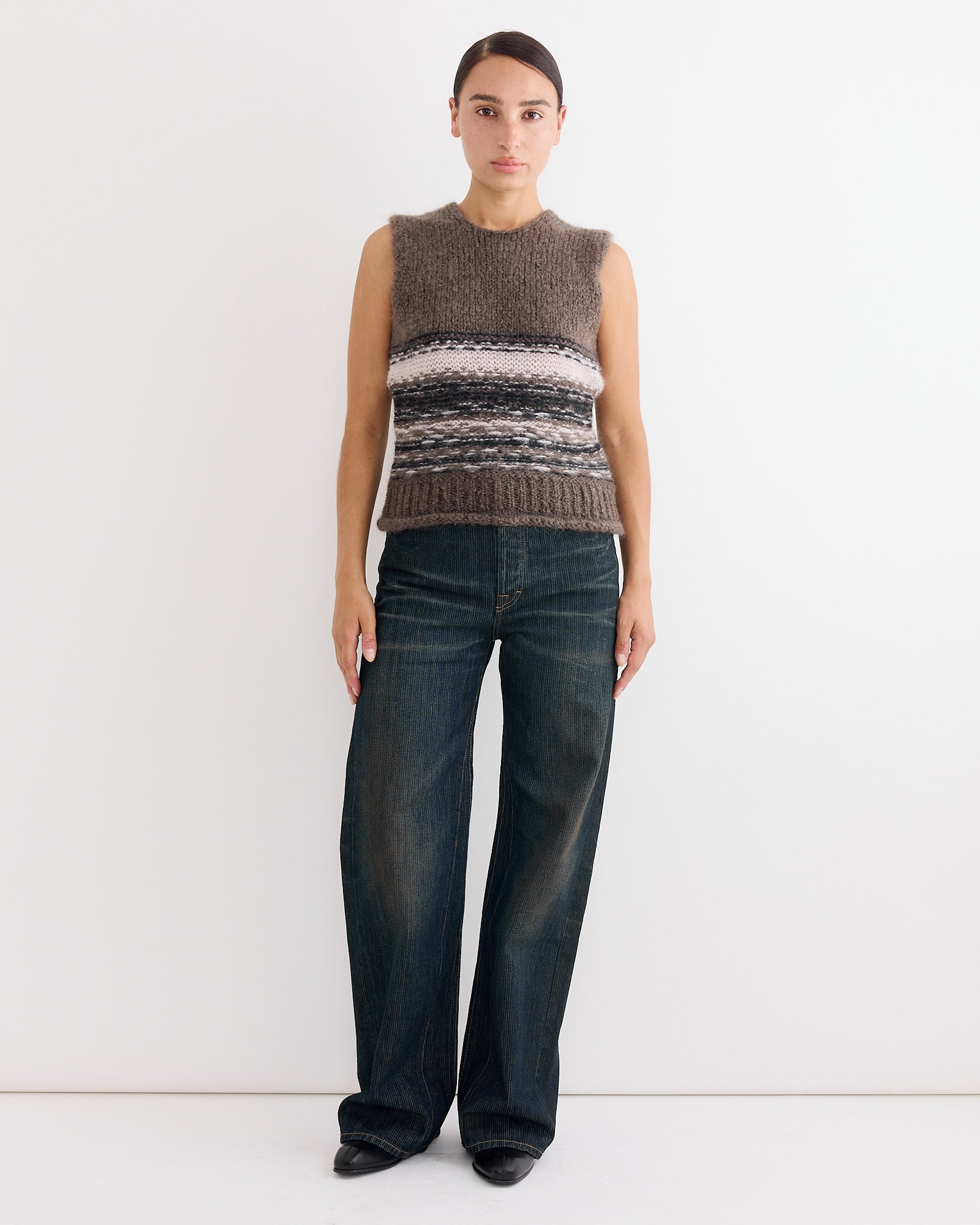 A person stands against a white wall wearing a sleeveless, textured knit sweater with horizontal stripes and wide-leg Our Legacy Treble Cut Jean in Clay Tint Chain Twill. Their dark hair is pulled back and they're wearing black pointed shoes.