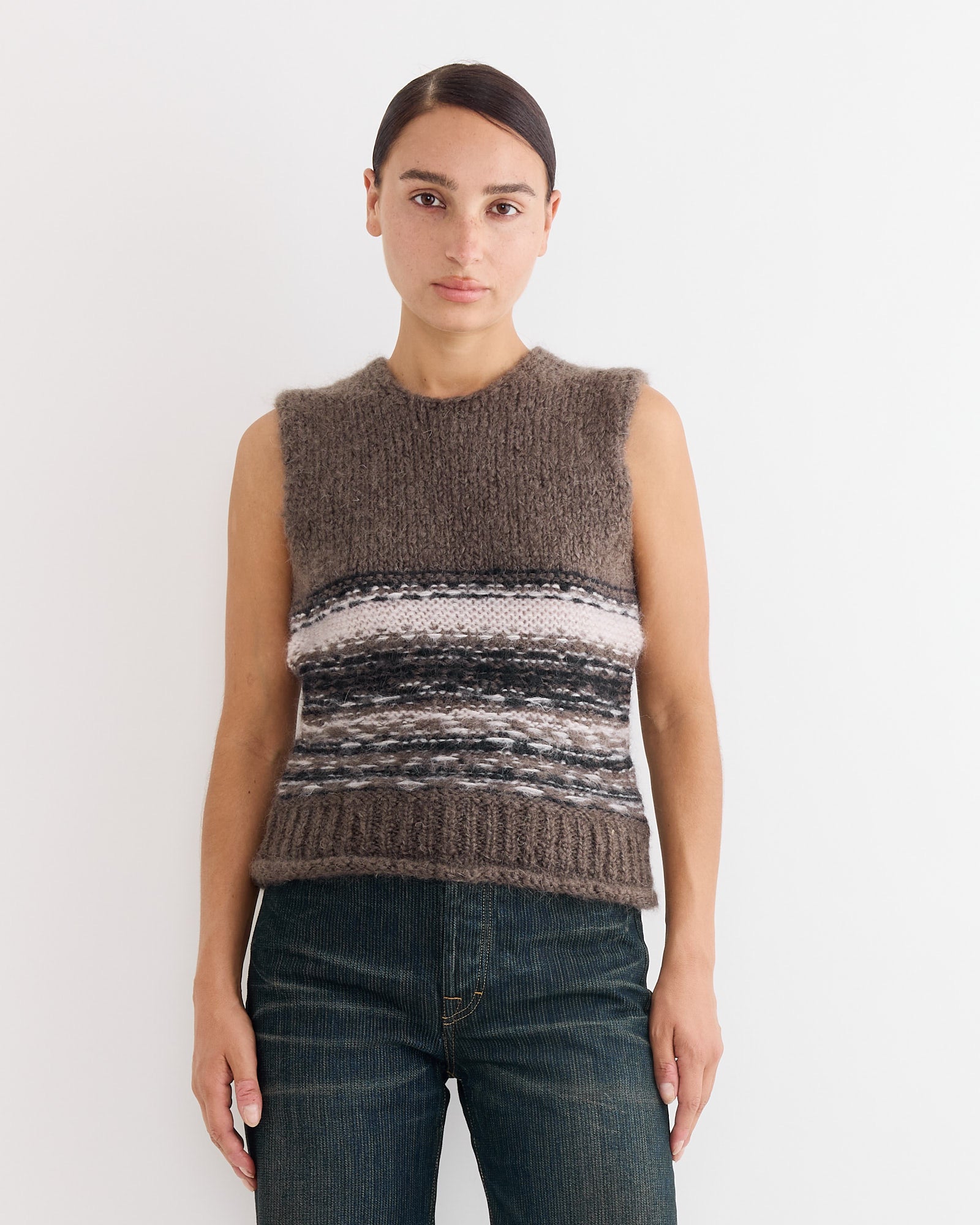 A person with straight dark hair, wearing the Our Legacy Pleaser Tank in Blur Fair Isle—featuring horizontal stripes—and blue jeans stands against a plain white background.