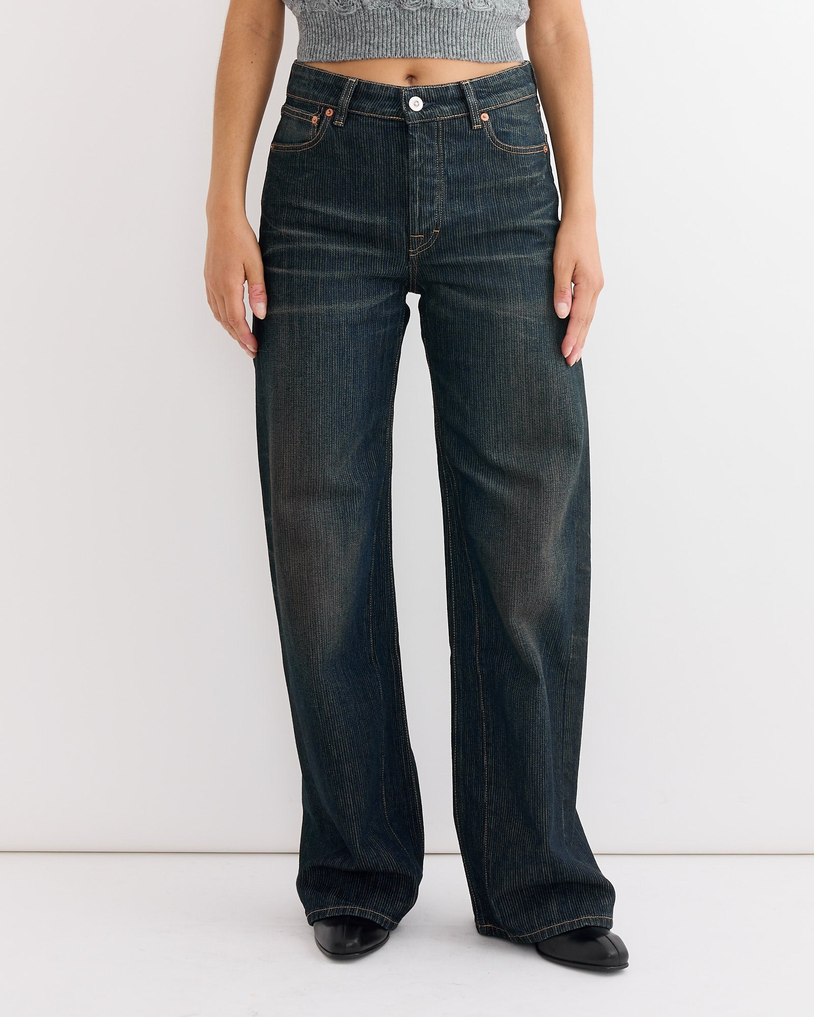 A person models the Our Legacy Treble Cut Jean in Clay Tint Chain Twill with a gray cropped top and black shoes, standing against a plain white background. The person’s head is not shown.