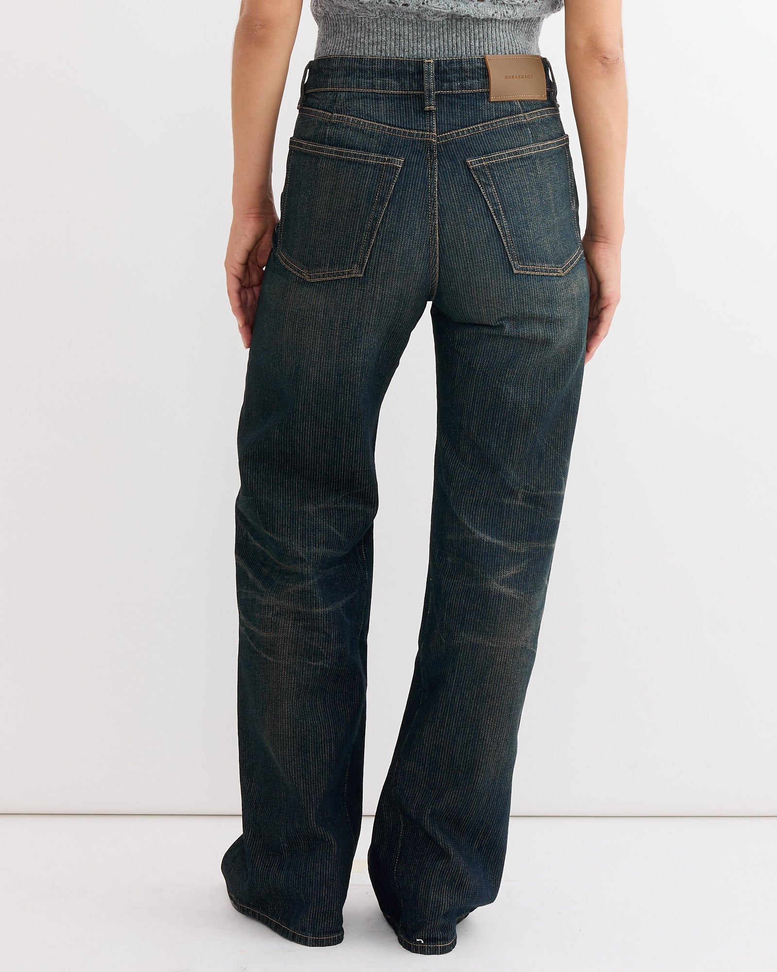 A person faces away, wearing Our Legacy Treble Cut Jean in Clay Tint Chain Twill—a high-waisted, straight-leg fit with visible seams and back pockets—paired with a gray textured top. The image highlights the lower body and denim details.