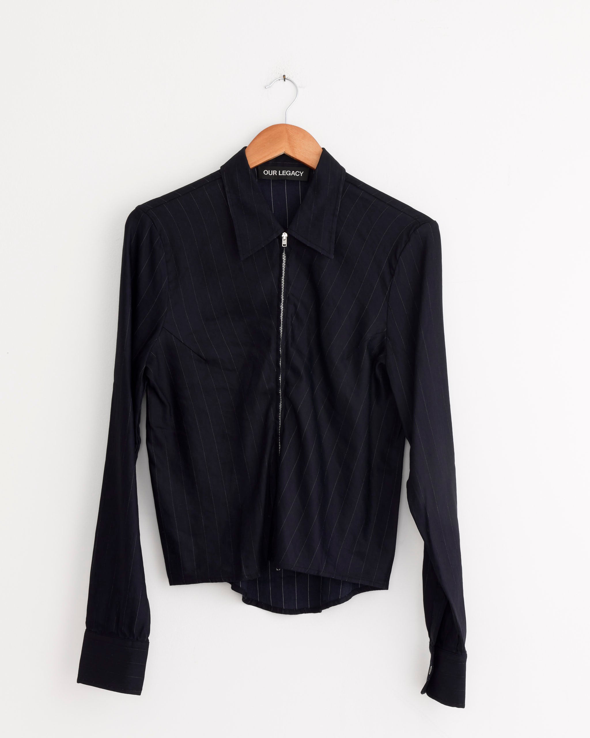 The Slim Zip Shirt in Night Covert Flex Pinstripe by Our Legacy, with a collar and front zipper, hangs on a wooden hanger against a white wall. Made from Italian pinstripe fabric, it features a regular slim fit and an OUR LEGACY tag.