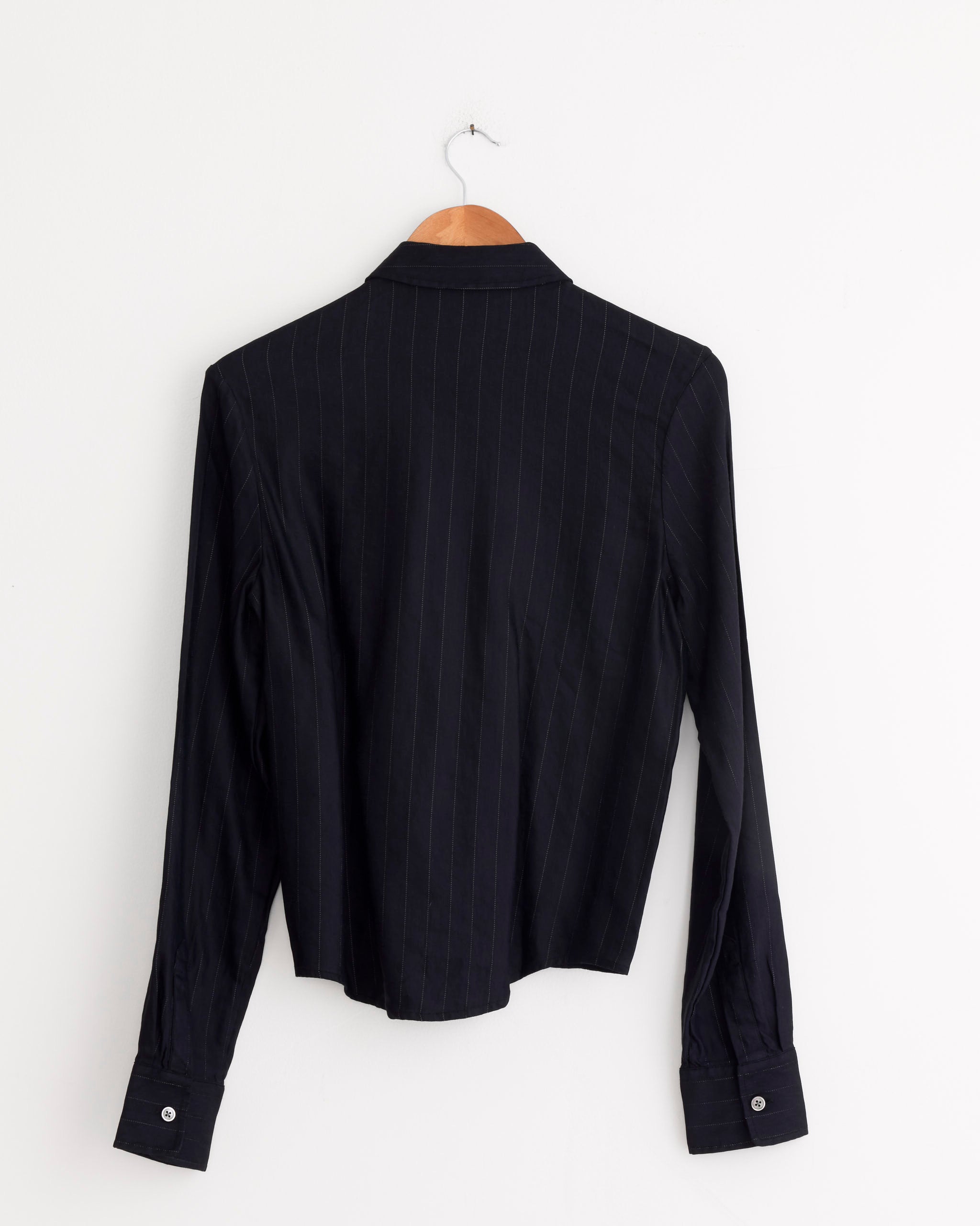 The Our Legacy Slim Zip Shirt in Night Covert Flex Pinstripe, featuring black pinstripe fabric, long sleeves, and a zip front, hangs on a wooden hanger against a plain white wall with the back view shown.