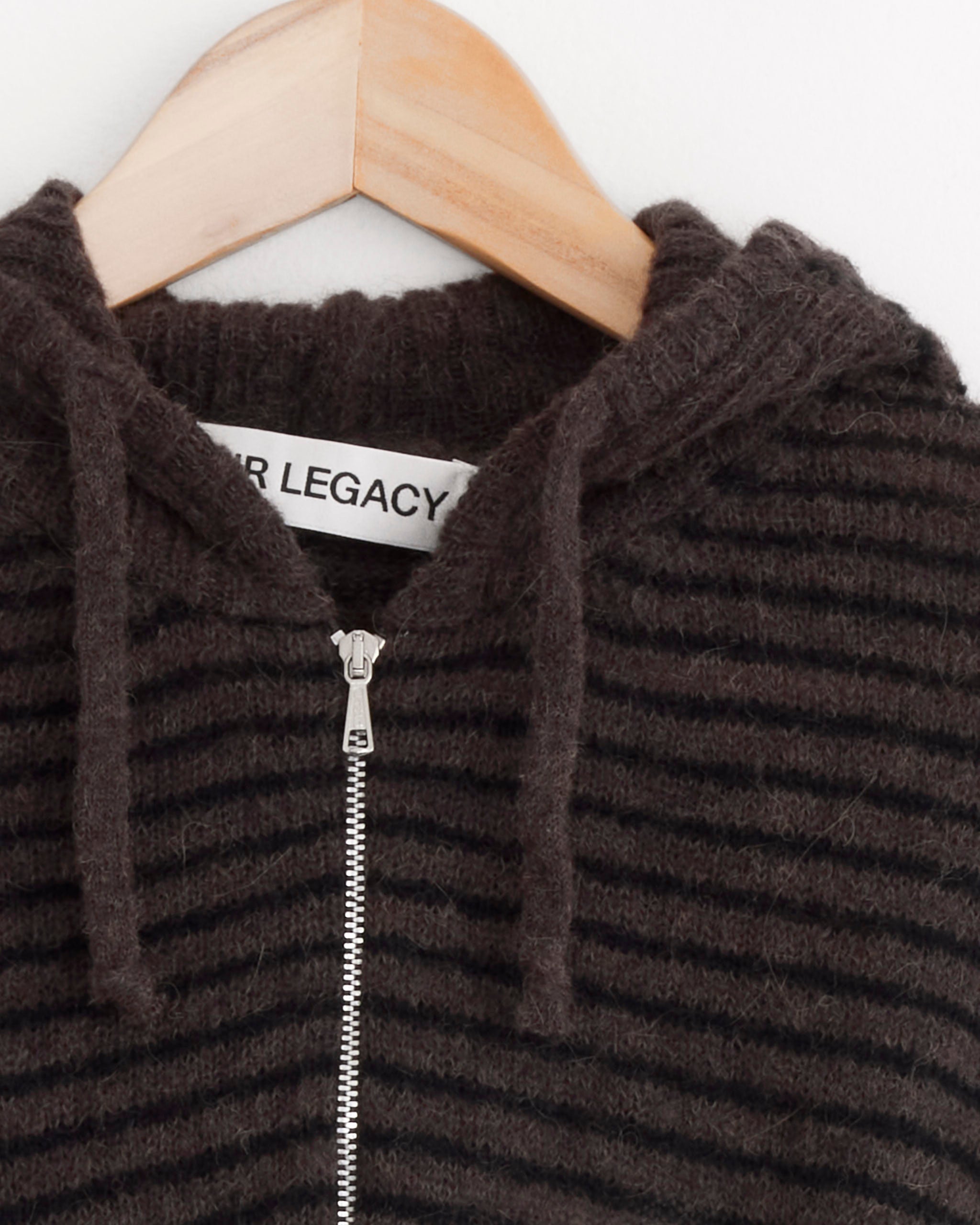 Close-up of the Our Legacy Compact Hood in Dirt Stripe Fuzzy Alpaca, an Italian yarn knit zip-up hoodie with a ribbed texture, visible zipper, hood cords, and partial “ER LEGACY” label, displayed on a wooden hanger.