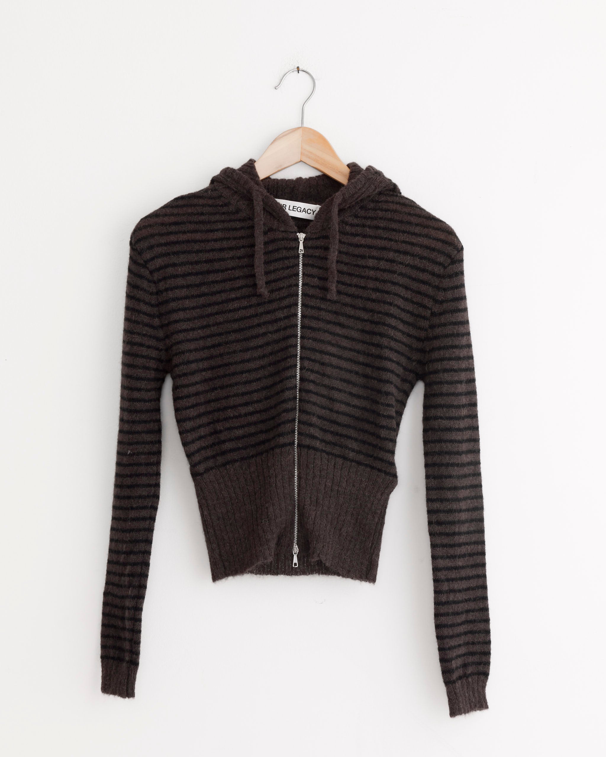 The Compact Hood in Dirt Stripe Fuzzy Alpaca by Our Legacy, a cropped dark brown zip-up hoodie with black stripes, crafted from luxurious Italian yarn knit, hangs on a wooden hanger against a plain white wall.