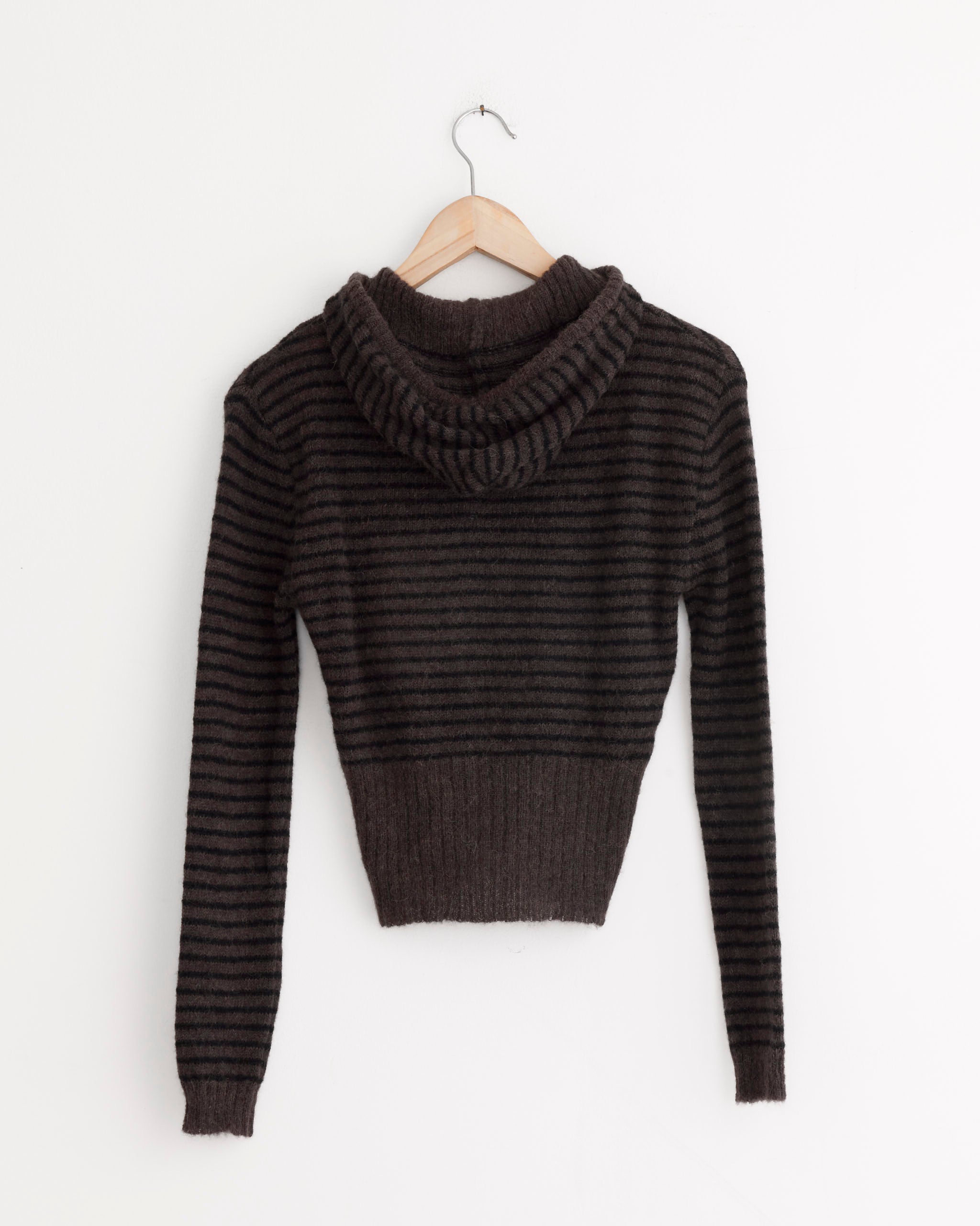 The Our Legacy Compact Hood in Dirt Stripe Fuzzy Alpaca, a dark brown, hooded, long-sleeve sweater with black stripes and ribbed hem and cuffs, hangs on a wooden hanger against a plain white wall.