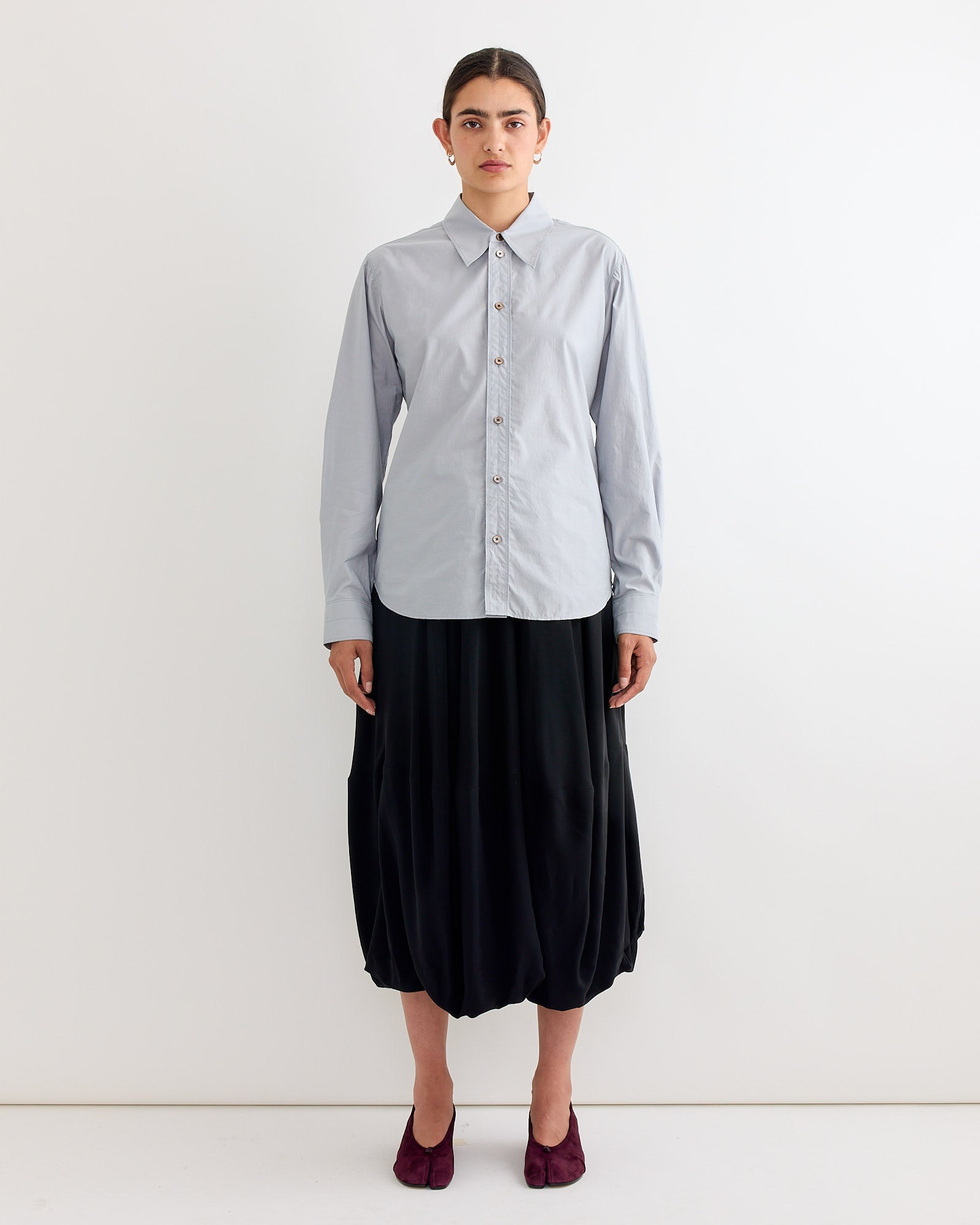 A person with dark hair pulled back and a neutral expression stands against a white background wearing the Lemaire Pointed Collar Shirt in Mist, paired with a loose black midi skirt and dark purple shoes.