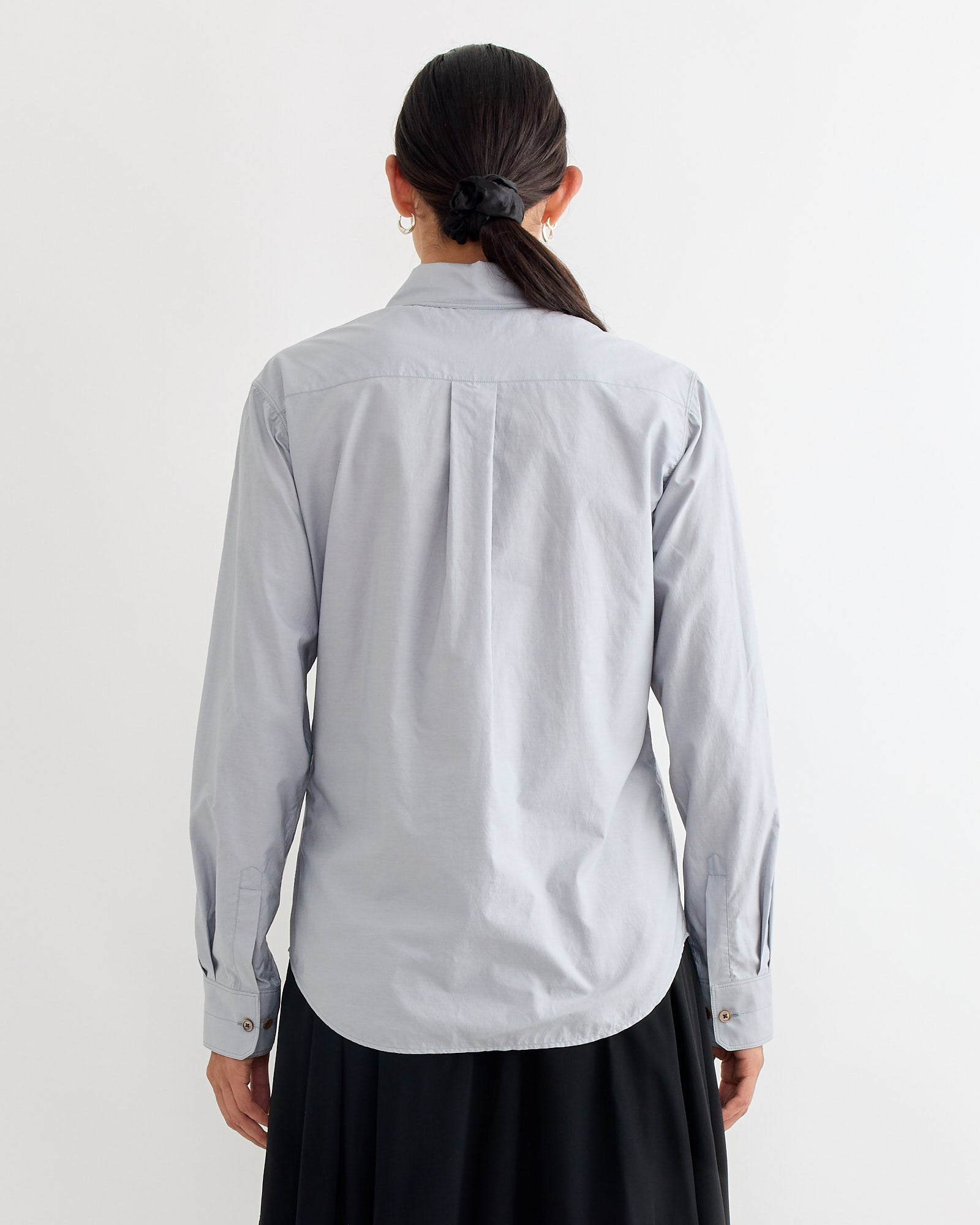 A person with long dark hair in a low ponytail is shown from the back wearing the Lemaire Pointed Collar Shirt in Mist and a black skirt, set against a plain white background.