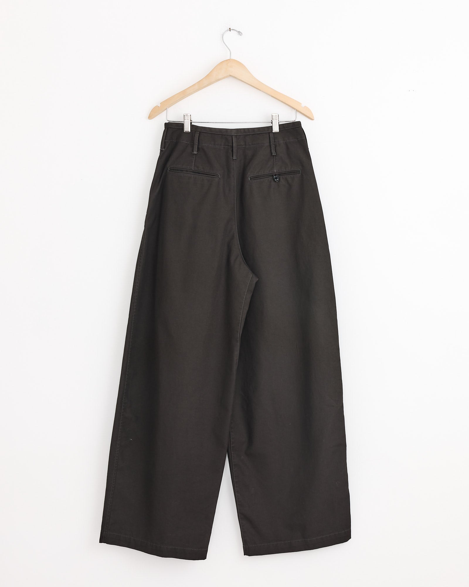 The Lemaire Large Chino in Espresso, crafted from Japanese cotton twill, hangs on a wooden hanger against a white wall, shown from the back with an oversized silhouette, two rear pockets, and a single button on the right pocket.