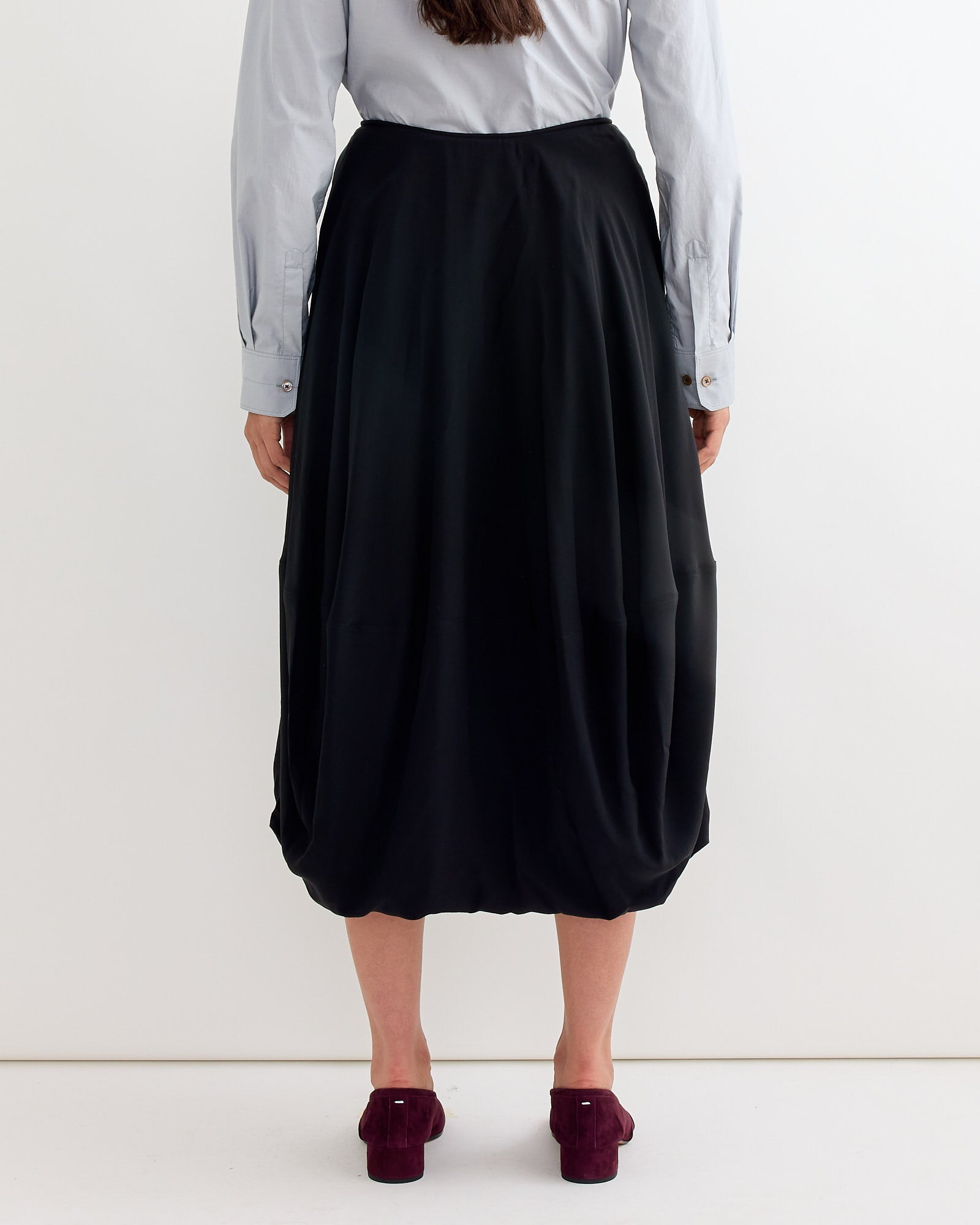A person stands facing away, wearing a light blue long-sleeve shirt, the Lemaire Balloon Skirt in Black, and purple heeled shoes, against a plain white background.