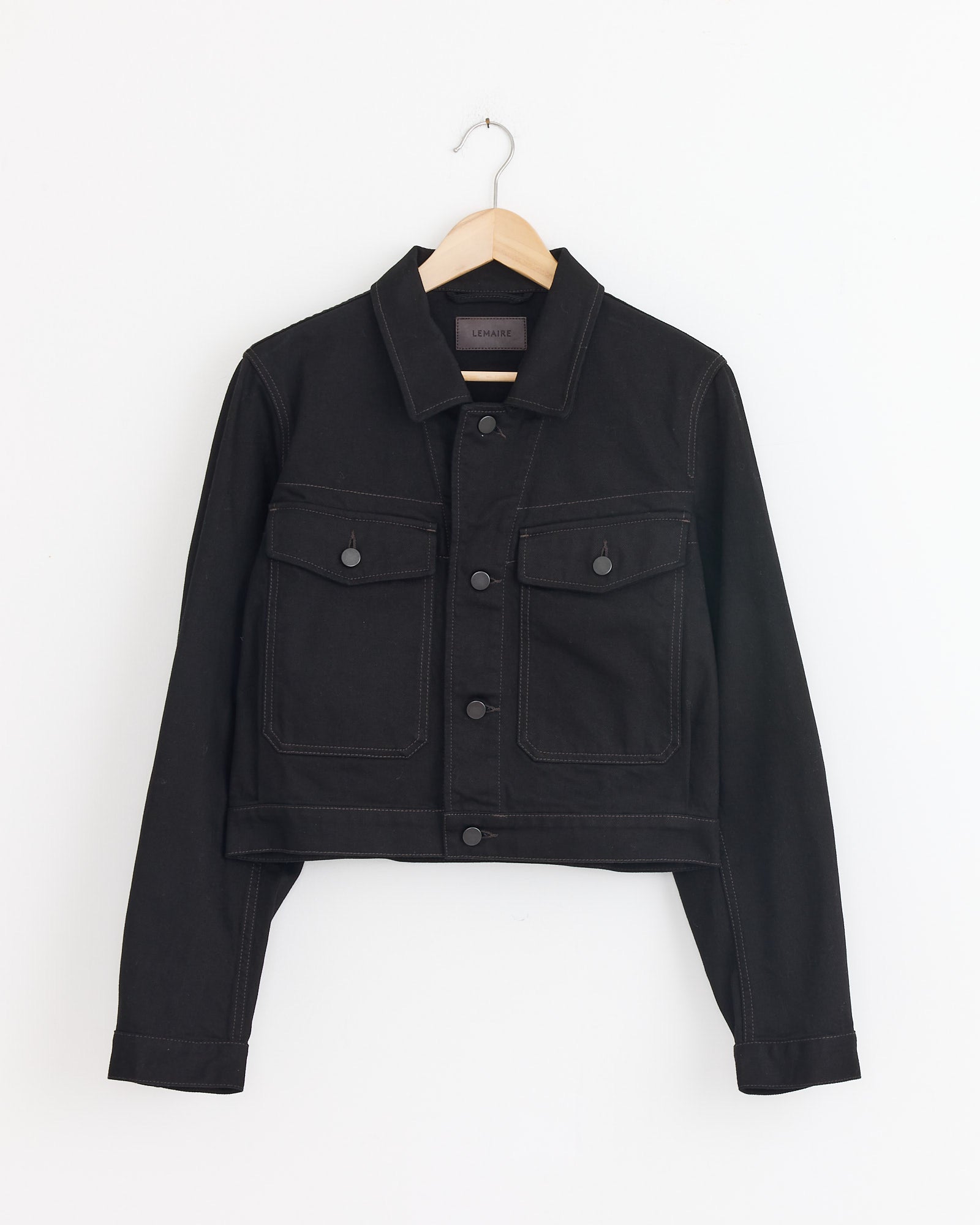 The Lemaire Cropped Blouson in Black is a workwear-inspired denim jacket featuring front buttons and two chest pockets, shown hanging on a wooden hanger against a white background.