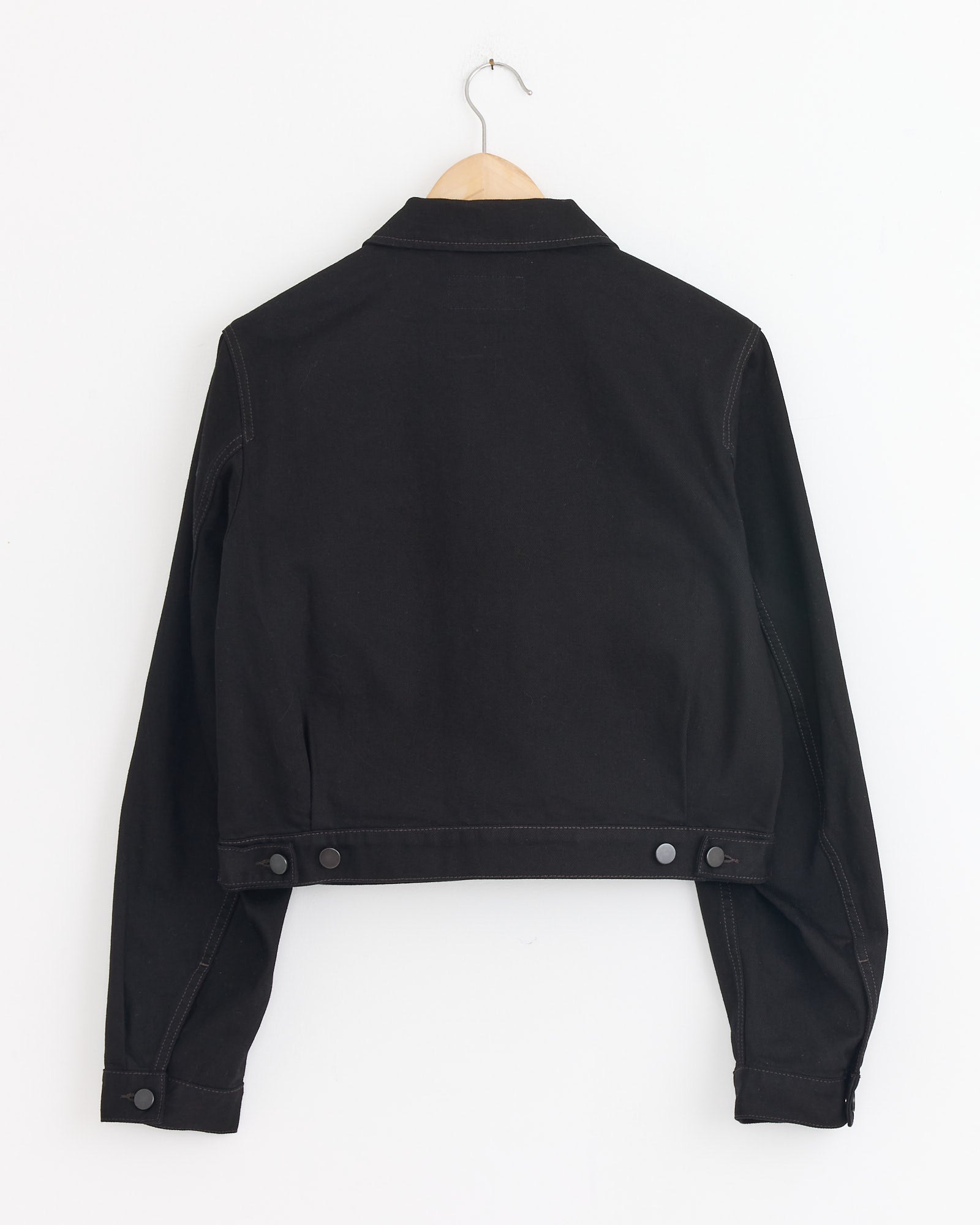 The Lemaire Cropped Blouson in Black, shown from the back on a wooden hanger against a white wall, features metal buttons on the waistband and cuffs for a workwear-inspired look.