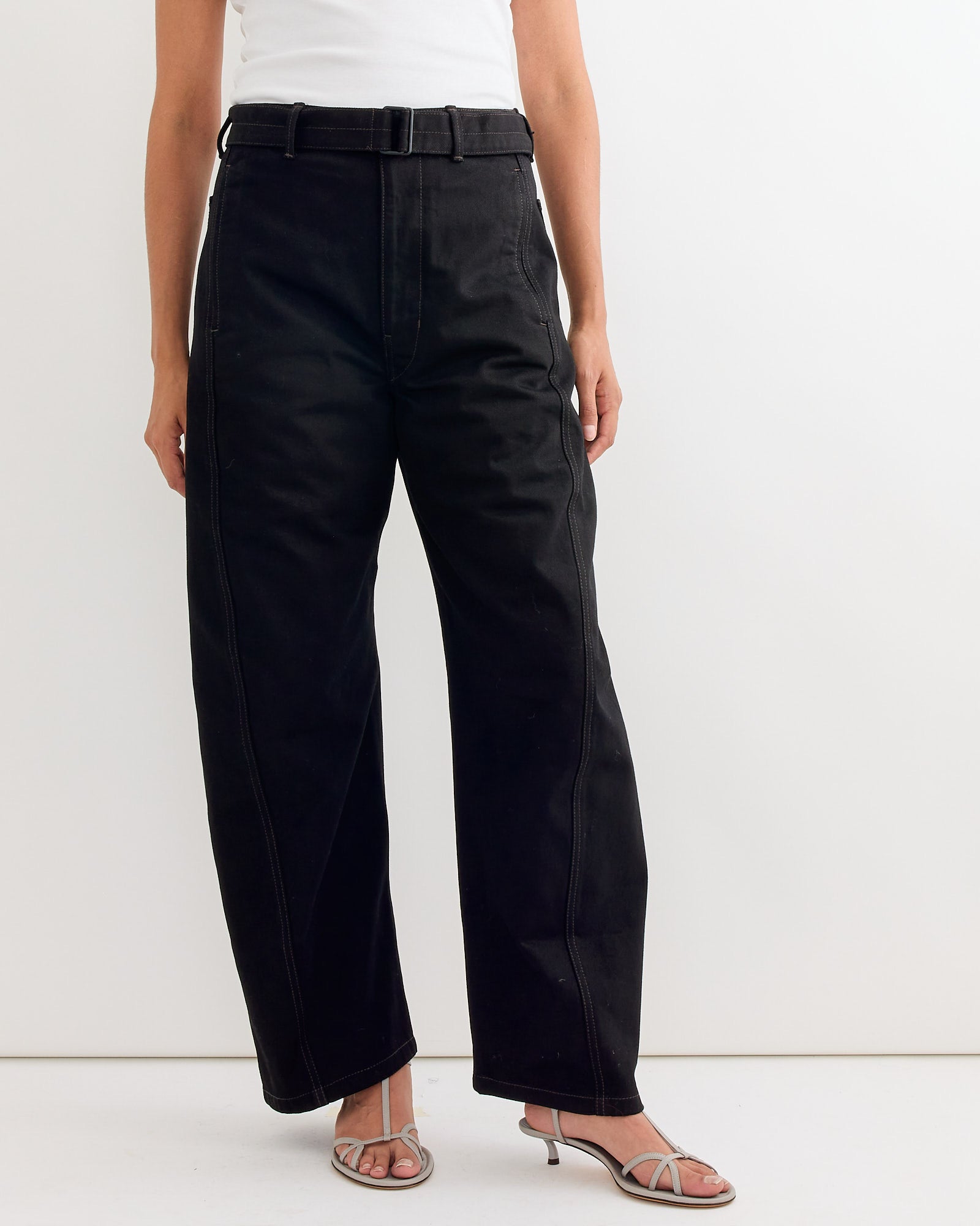 A person stands against a plain white background, shown from shoulders down, wearing Lemaire's Twisted Belted Pant in Black with a white sleeveless top and open-toe heeled sandals.