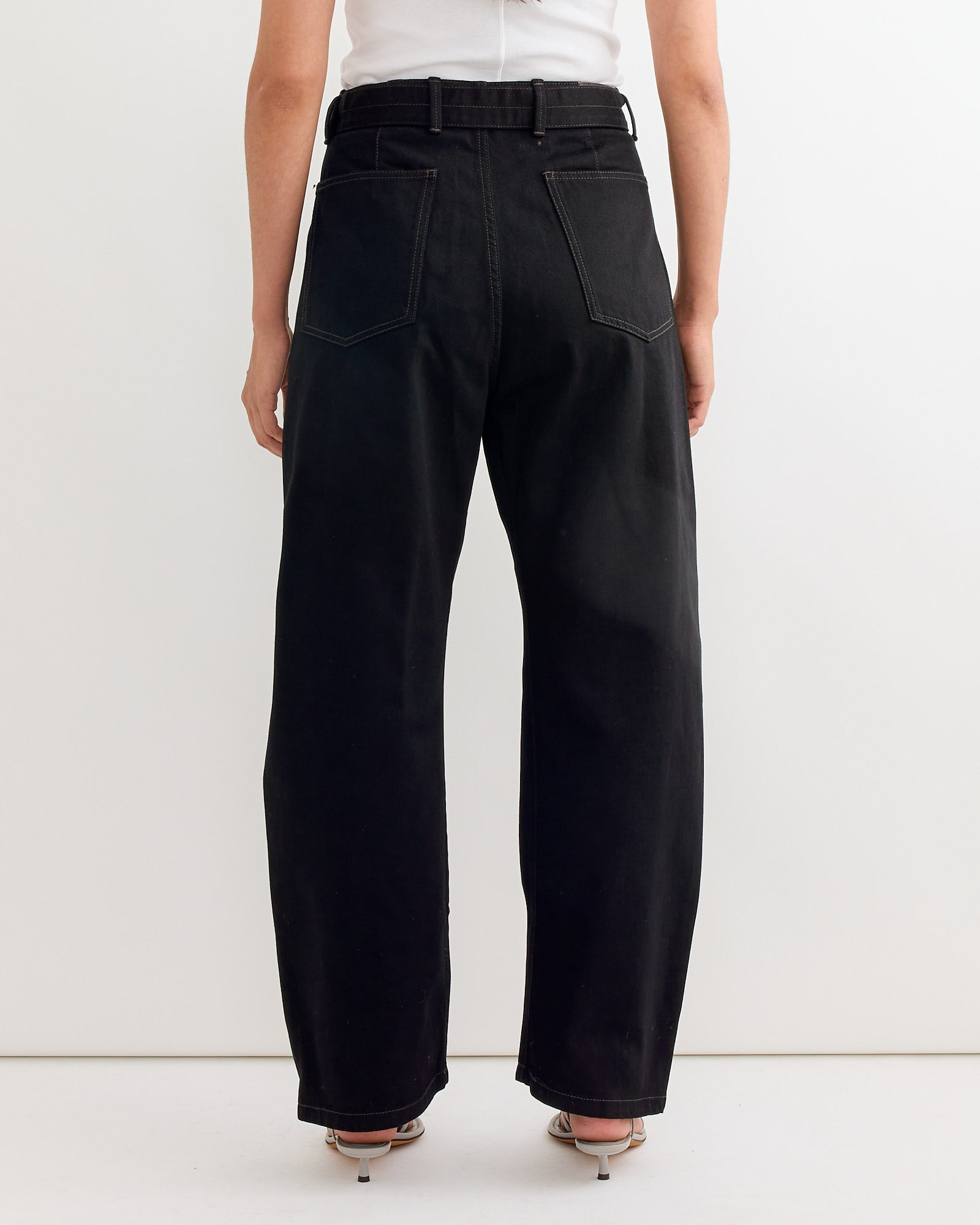 A person wearing Lemaire’s Twisted Belted Pant in Black, high-waisted and loose-fitting, styled with heeled sandals, shown from the back against a plain white background.