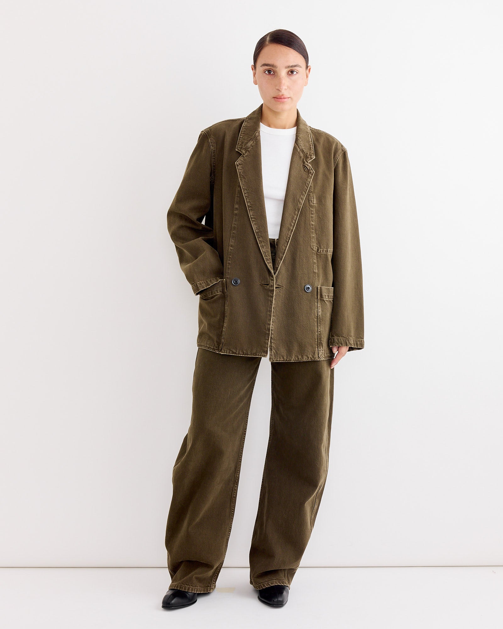 A person wears Lemaire's Workwear Blazer in Moss Green with matching wide-leg pants, a white shirt, and black shoes. The relaxed fit is complemented by dark hair pulled back and a neutral expression, set against a white wall.