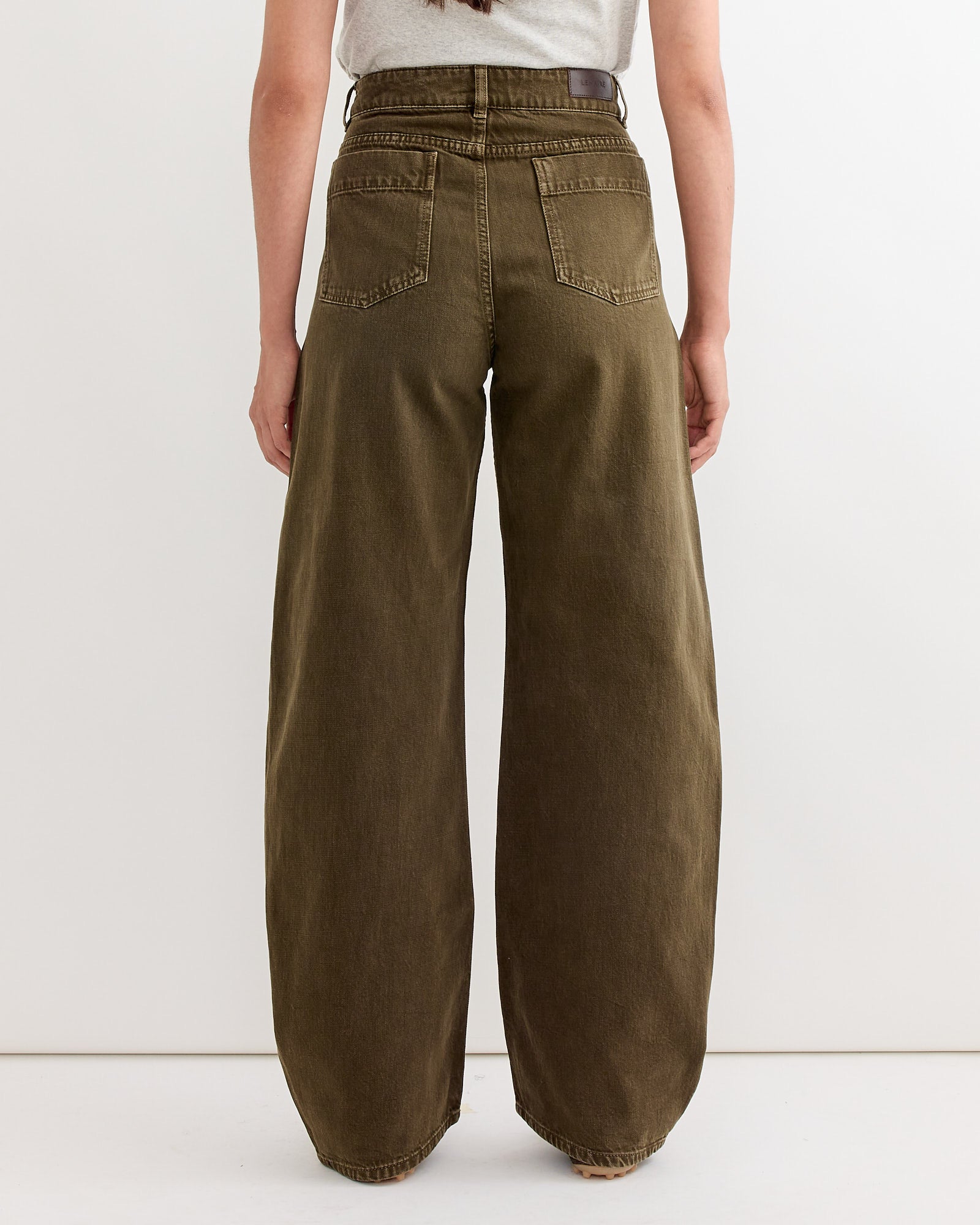 A person faces away from the camera, wearing Lemaire's High Waisted Curved Pant in Moss Green and a tucked-in light gray shirt, set against a plain white background.