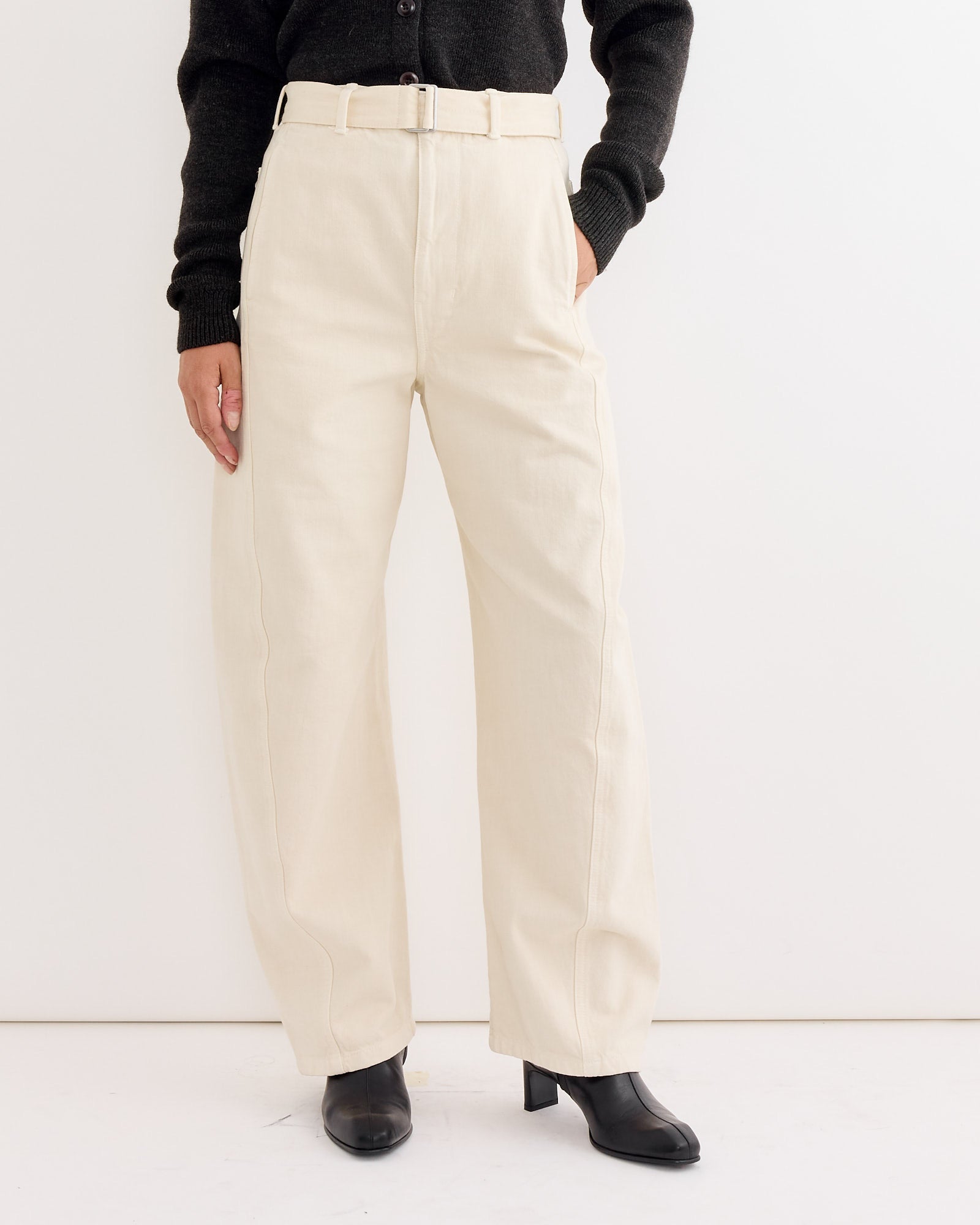A person in Lemaire's Twisted Belted Pant in Snow Clay, a black long-sleeve top, and black ankle boots stands against a plain white background, highlighting the pant's refined unisex design.