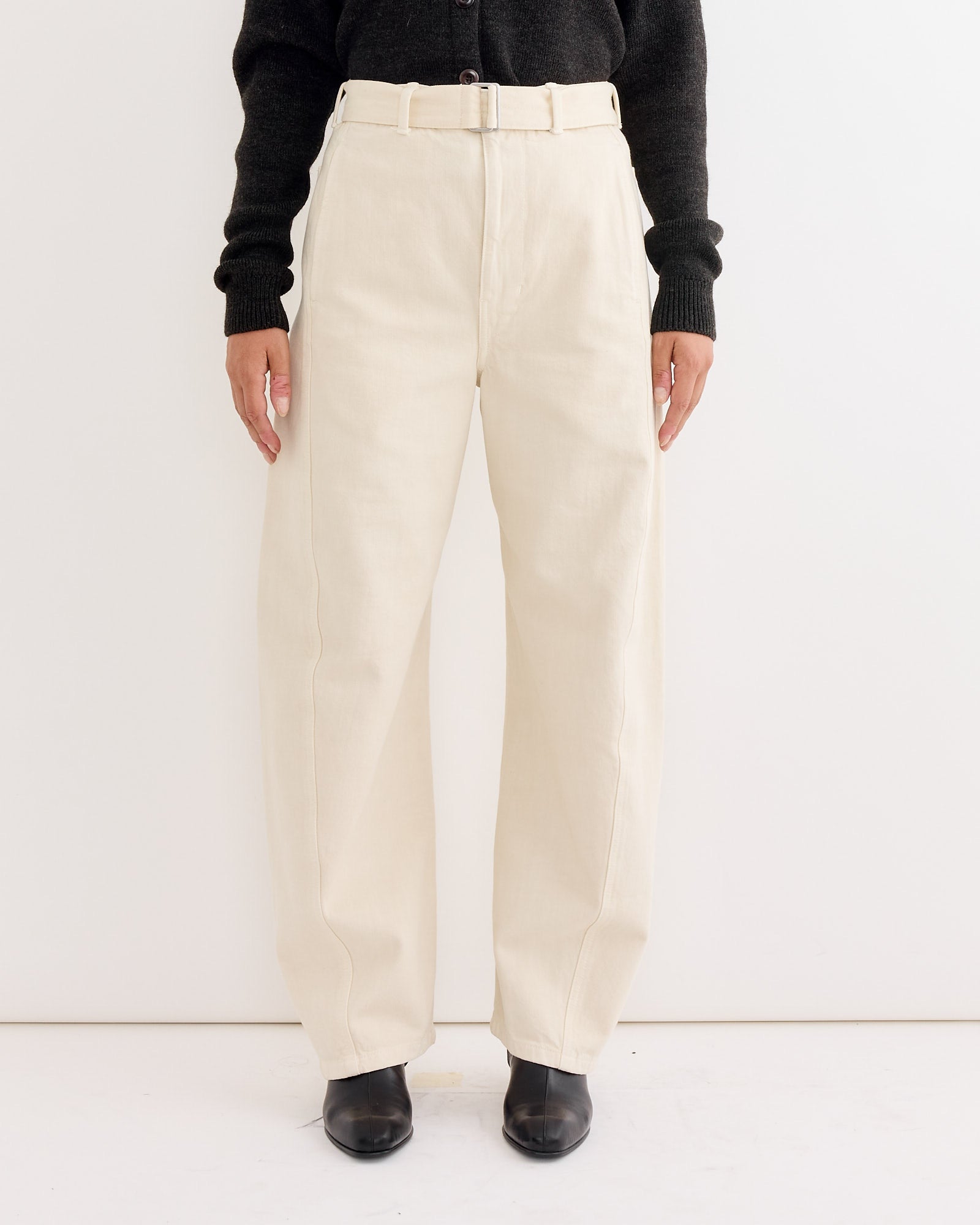 A person wearing high-waisted, wide-leg Lemaire Twisted Belted Pant in Snow Clay with a matching belt, paired with a black long-sleeve top and black shoes, stands against a plain white background.