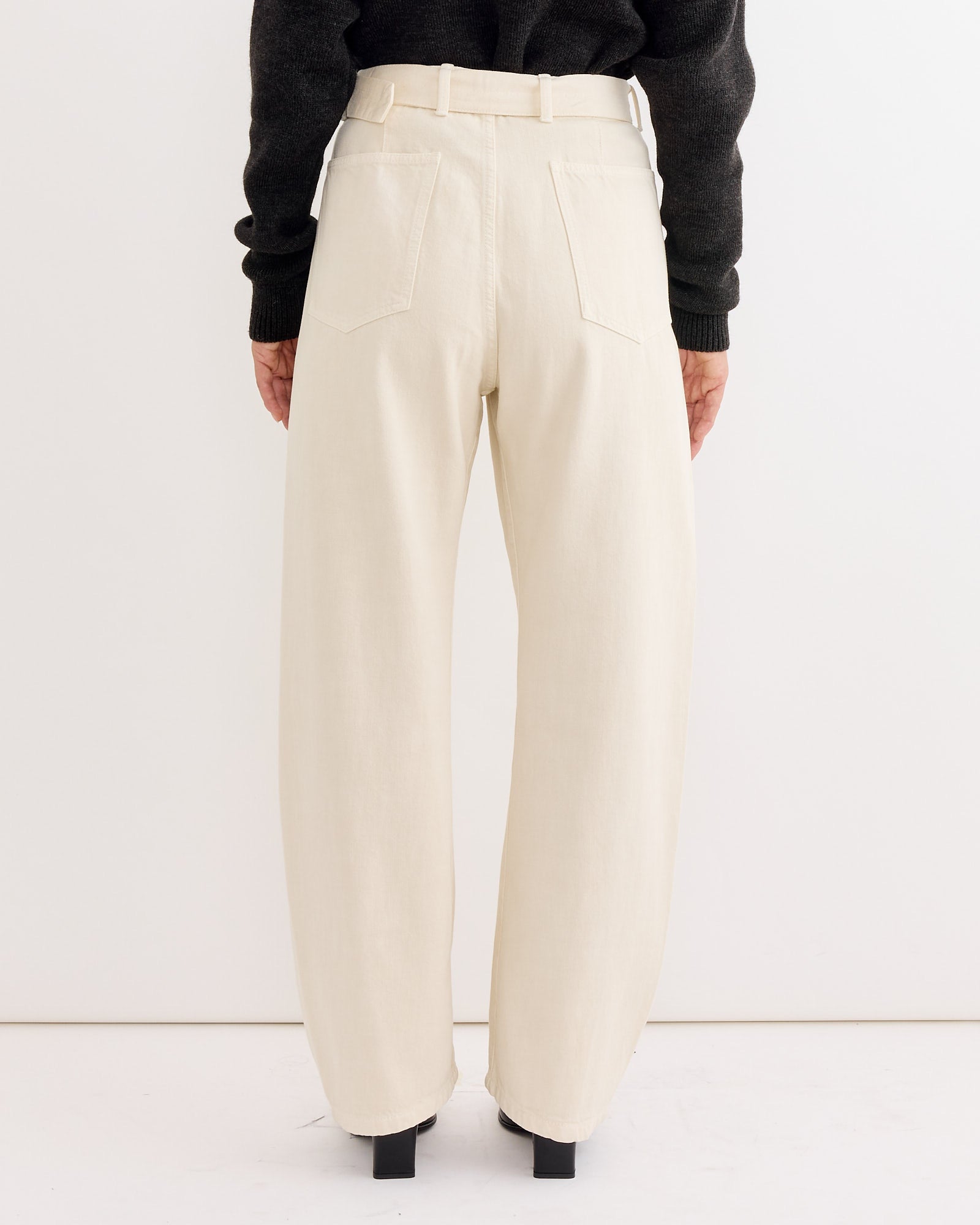 A person, seen from the back, stands against a plain white background wearing Lemaire’s Twisted Belted Pant in Snow Clay—high-waisted, loose-fitting pants with a unisex design—and a dark sweater.