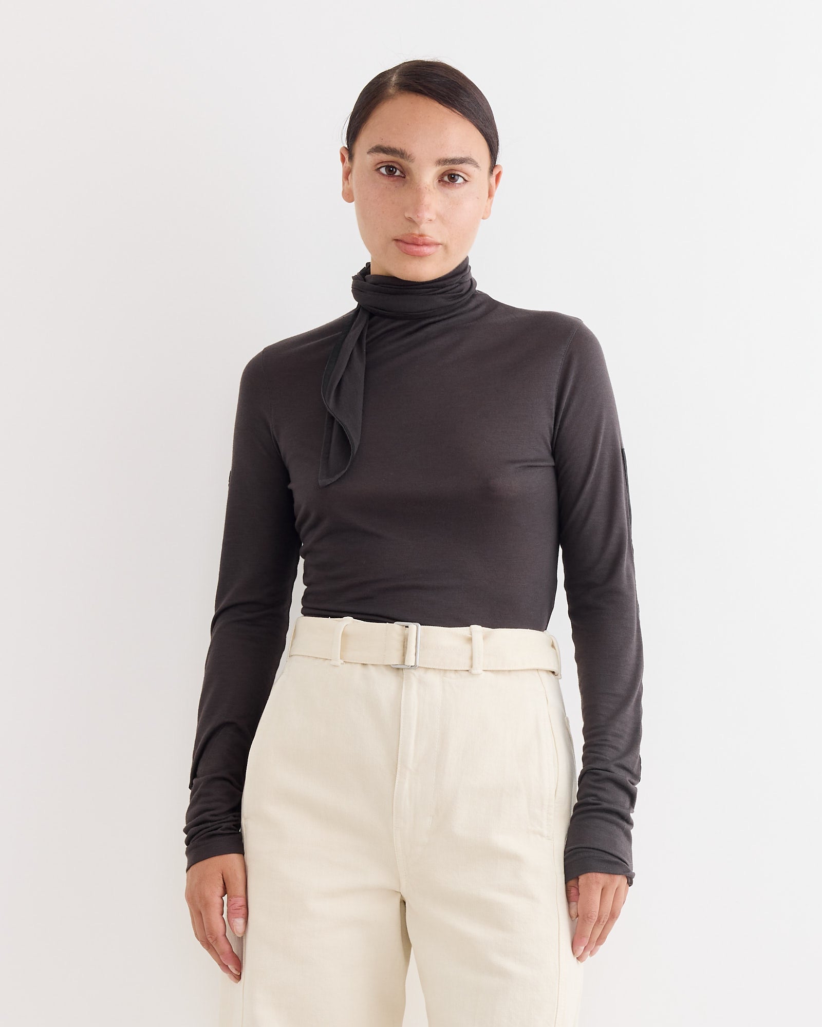 A person stands against a white background, wearing Lemaire’s fitted Foulard Patches Top in Coal with tie detail and high-waisted, light beige belted trousers.