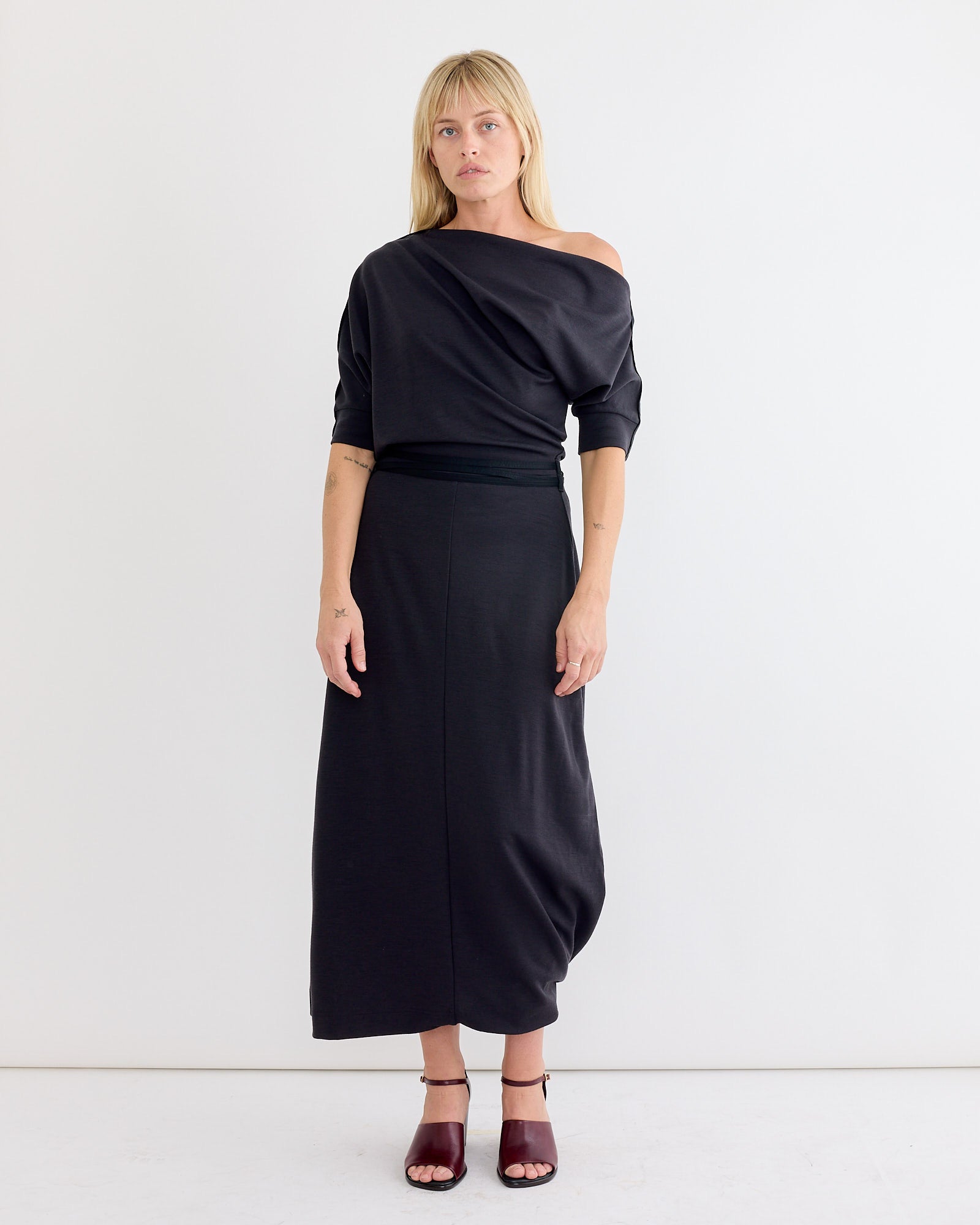 A person with blonde hair and bangs stands against a white background wearing the Lemaire Twisted Hem Dress in Anthracite, featuring an asymmetrical neckline and short sleeves, paired with maroon heeled sandals and a neutral expression.