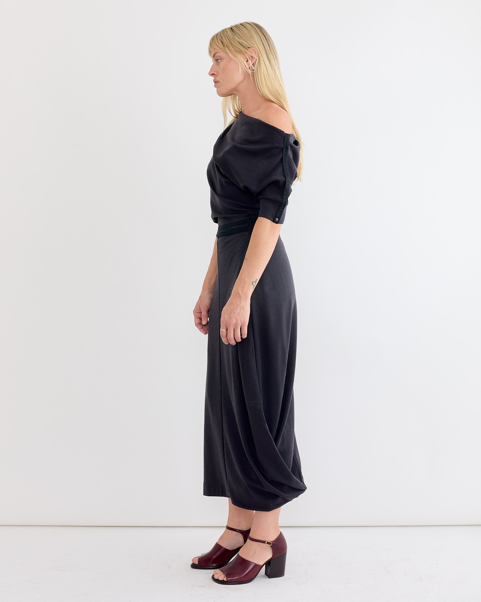 A woman with blonde hair stands sideways against a white background, wearing the Lemaire Twisted Hem Dress in Anthracite and maroon heeled sandals. Her arms are relaxed by her sides.