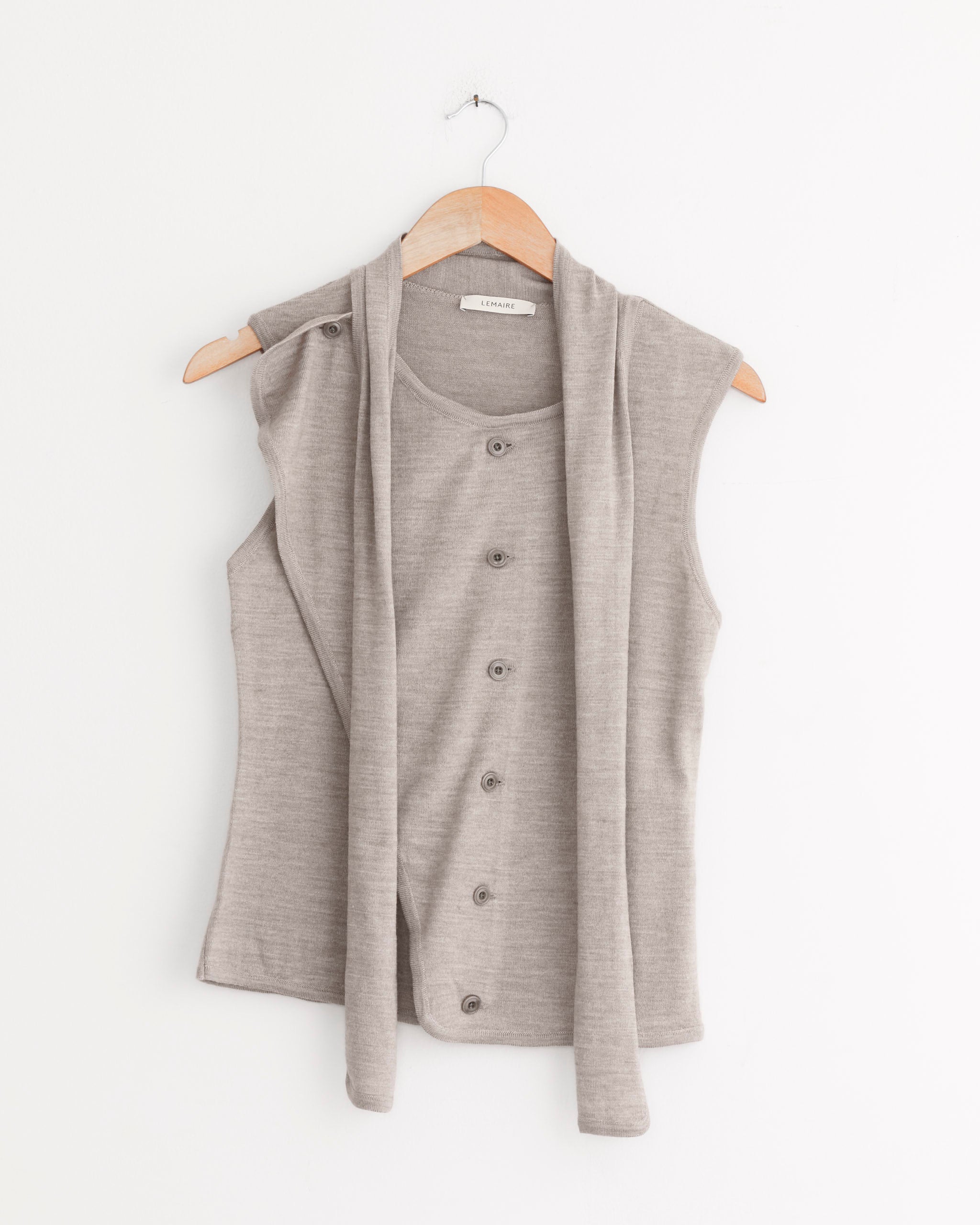 The Lemaire Asymmetrical Sleeveless Cardigan in Light Taupe, featuring a button-down front and matching long scarf, is displayed on a wooden hanger against a white wall.