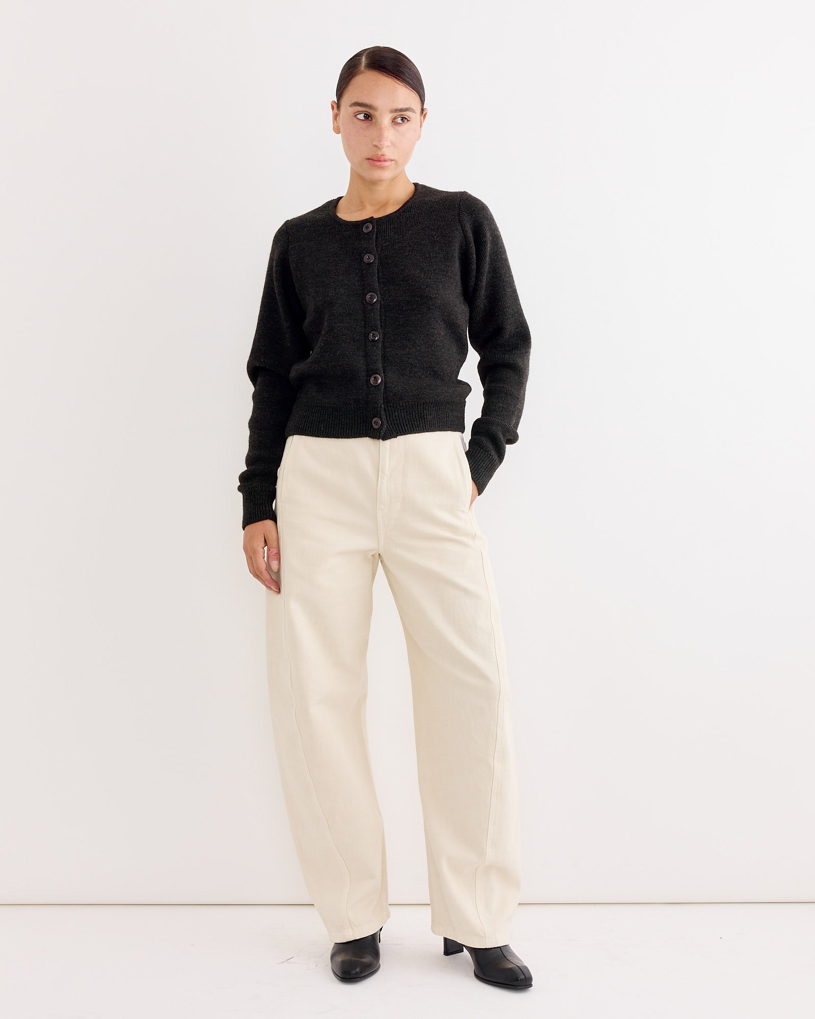 A woman with her hair pulled back and a neutral expression stands against a white wall, wearing the Lemaire Cardigan Dark in Espresso, cream wide-leg pants, and black shoes.