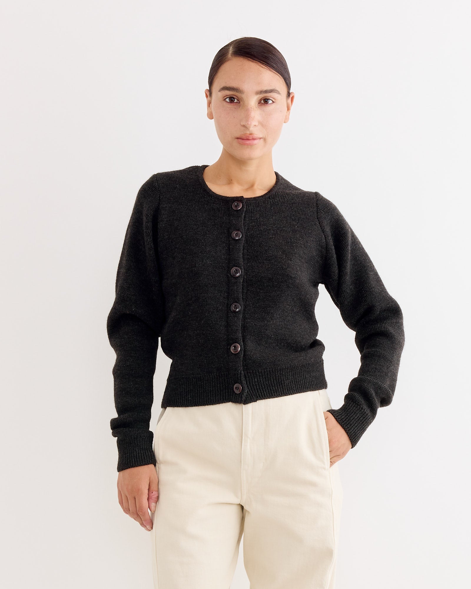 A person stands against a plain white background, wearing the Lemaire Cardigan Dark in Espresso and high-waisted light beige pants, with one hand in their pocket and a neutral expression.