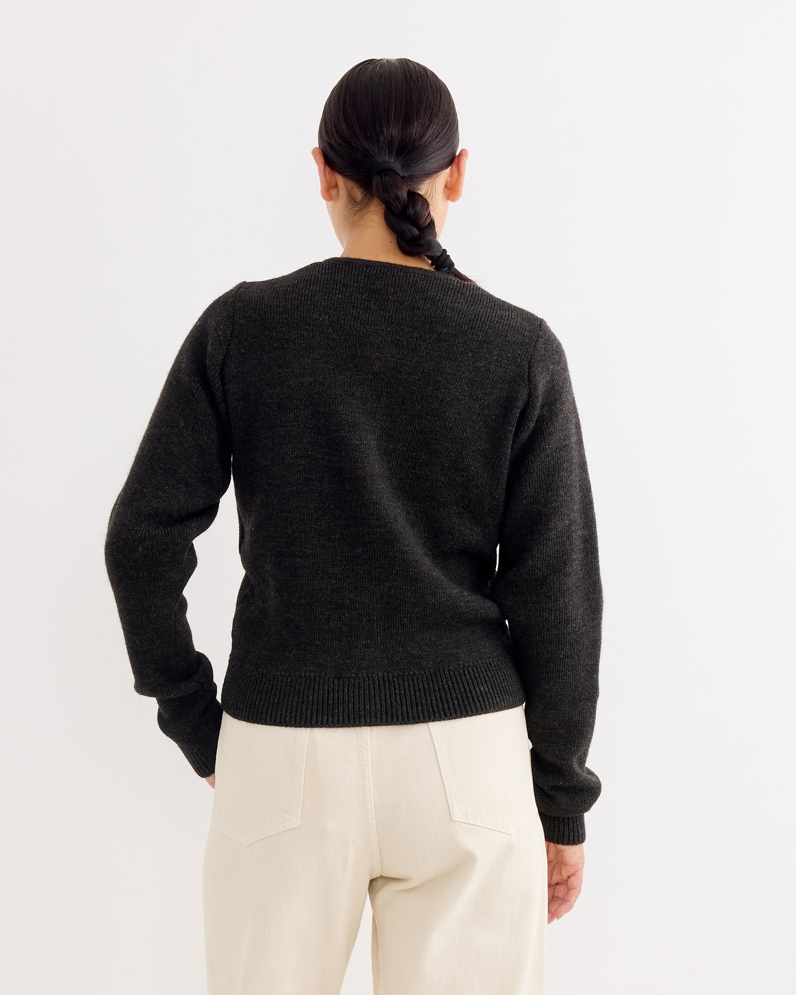 A person with dark, braided hair faces away, wearing the Lemaire Cardigan Dark in Espresso paired with cream pants, set against a plain light background.