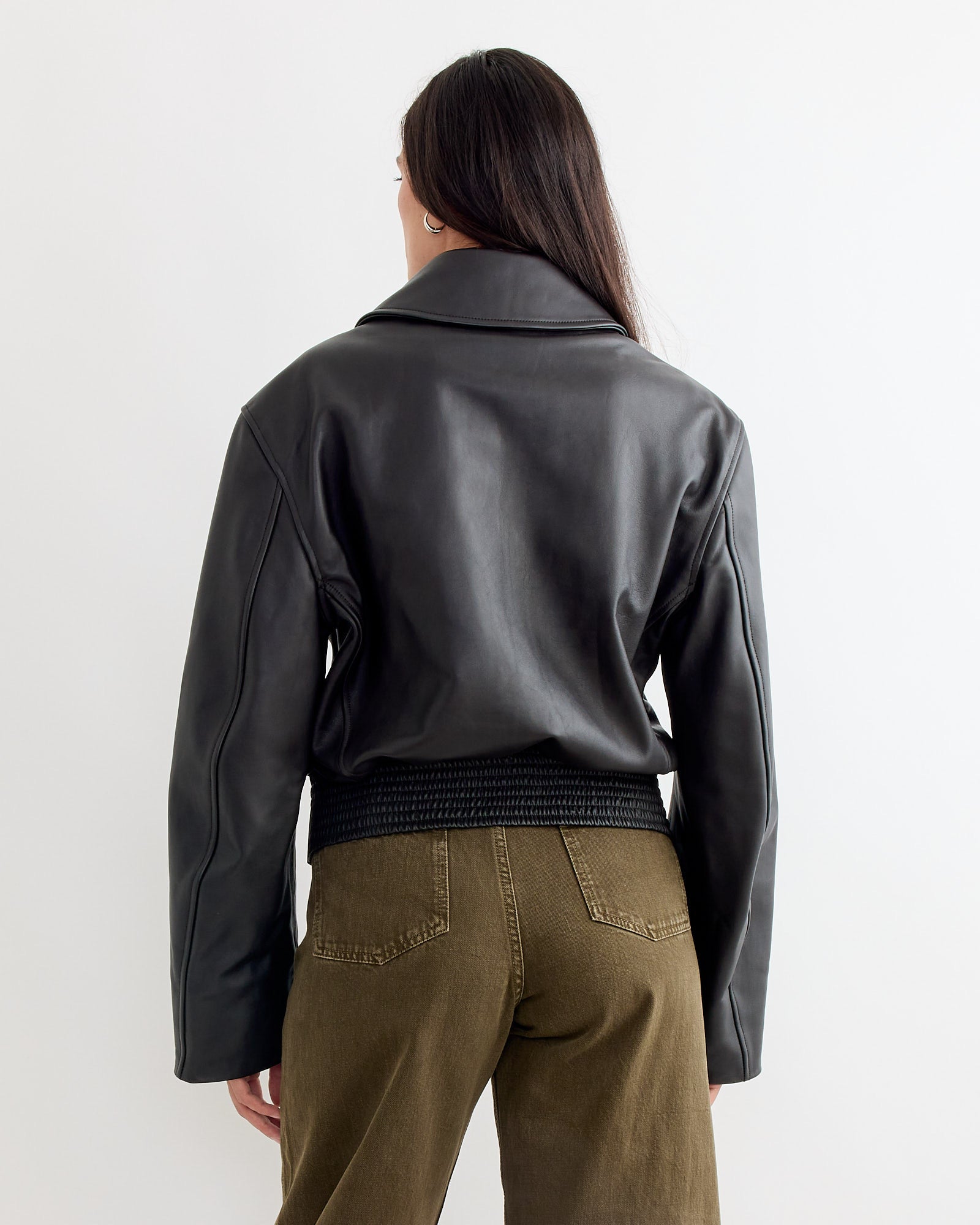 A person with long dark hair is seen from behind, wearing the Lemaire Boxy Leather Blouson in Moka and olive green pants, standing against a plain white background.